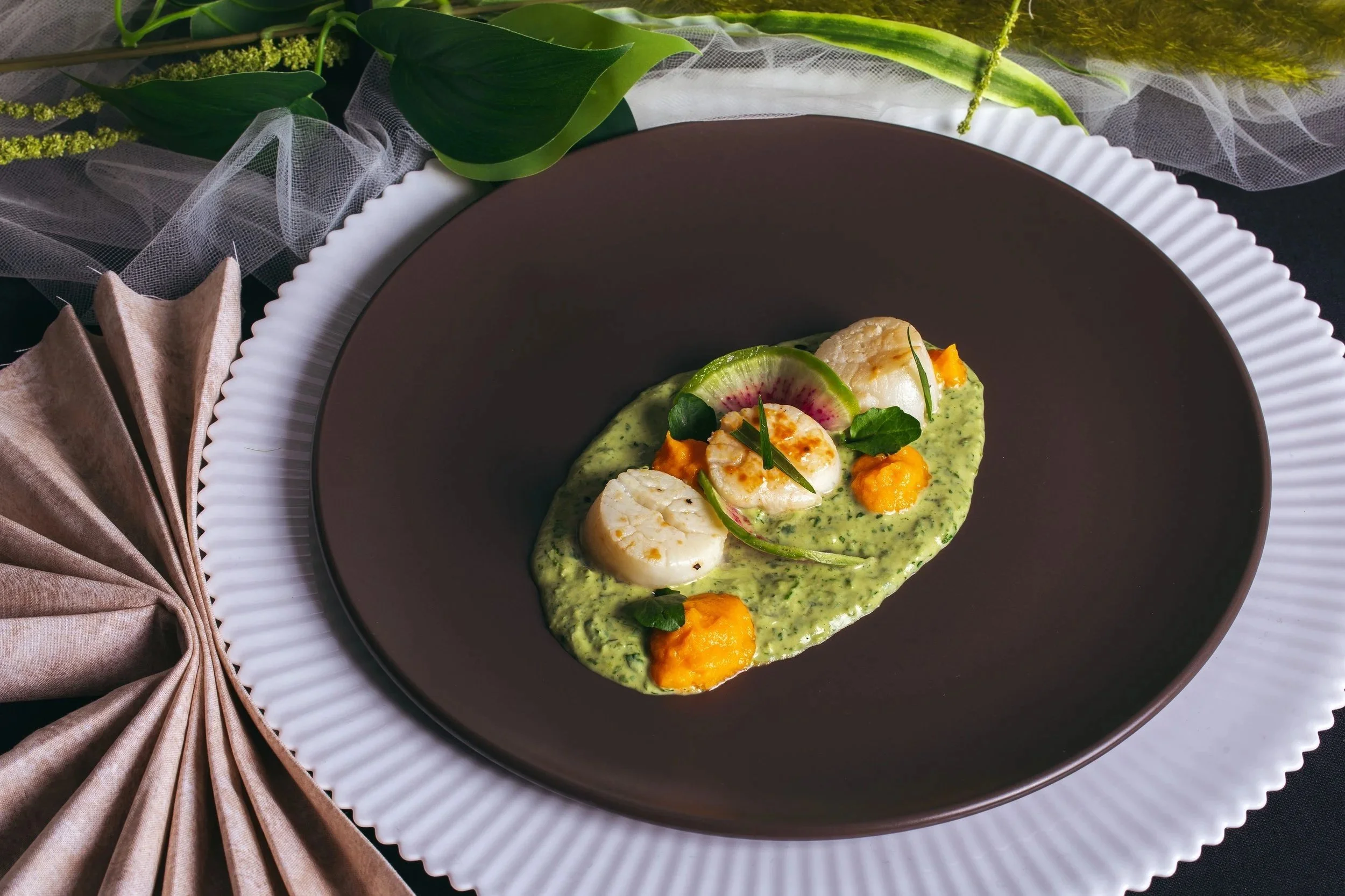 A gourmet dish with scallops atop a green sauce, garnished with slices of radish, small orange dollops, and fresh herbs, presented on a modern black and white plate.