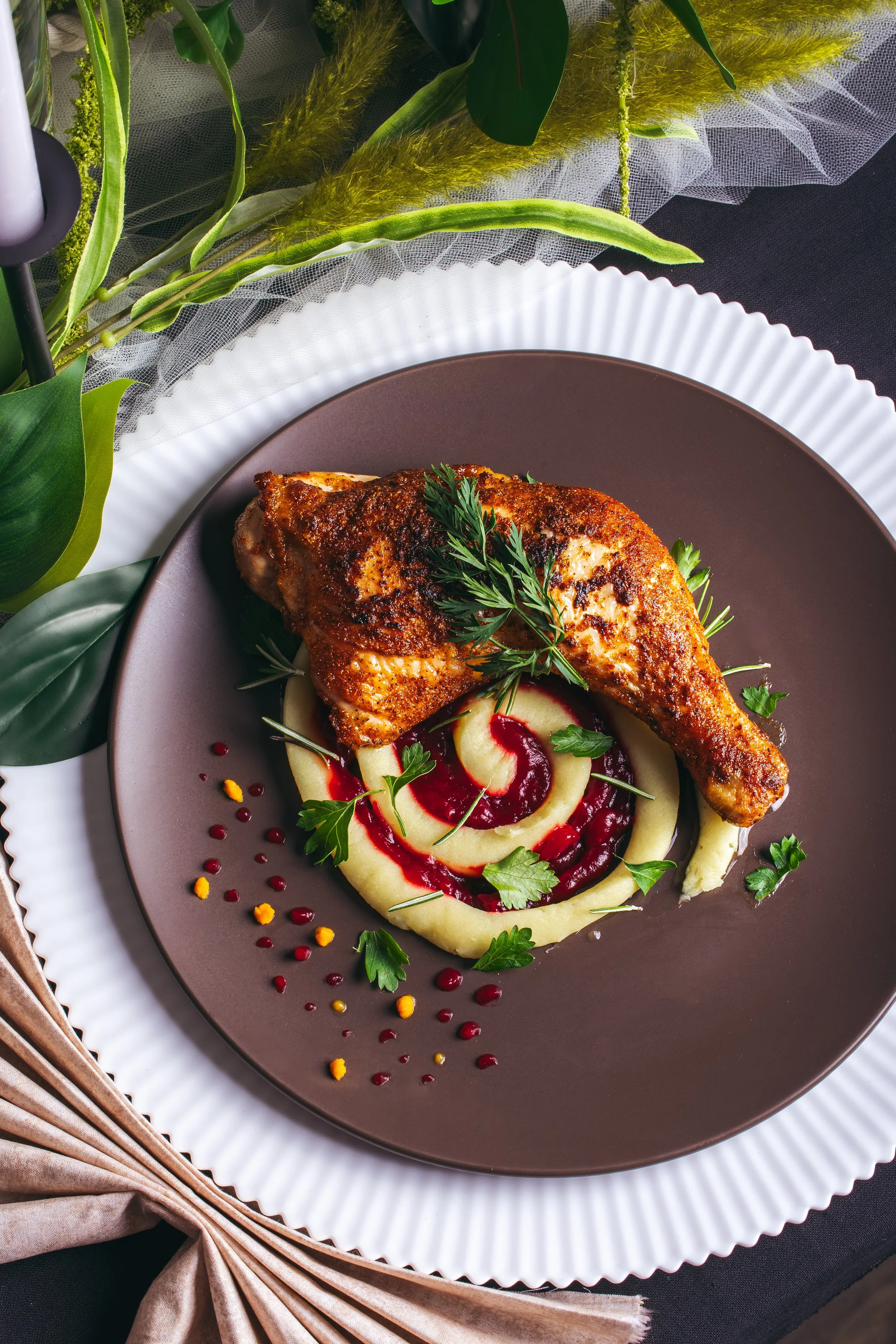 A plate with a roasted chicken leg, garnished with herbs, served on a bed of mashed potatoes with red sauce drizzle and parsley, on a decorative brown and white plates.