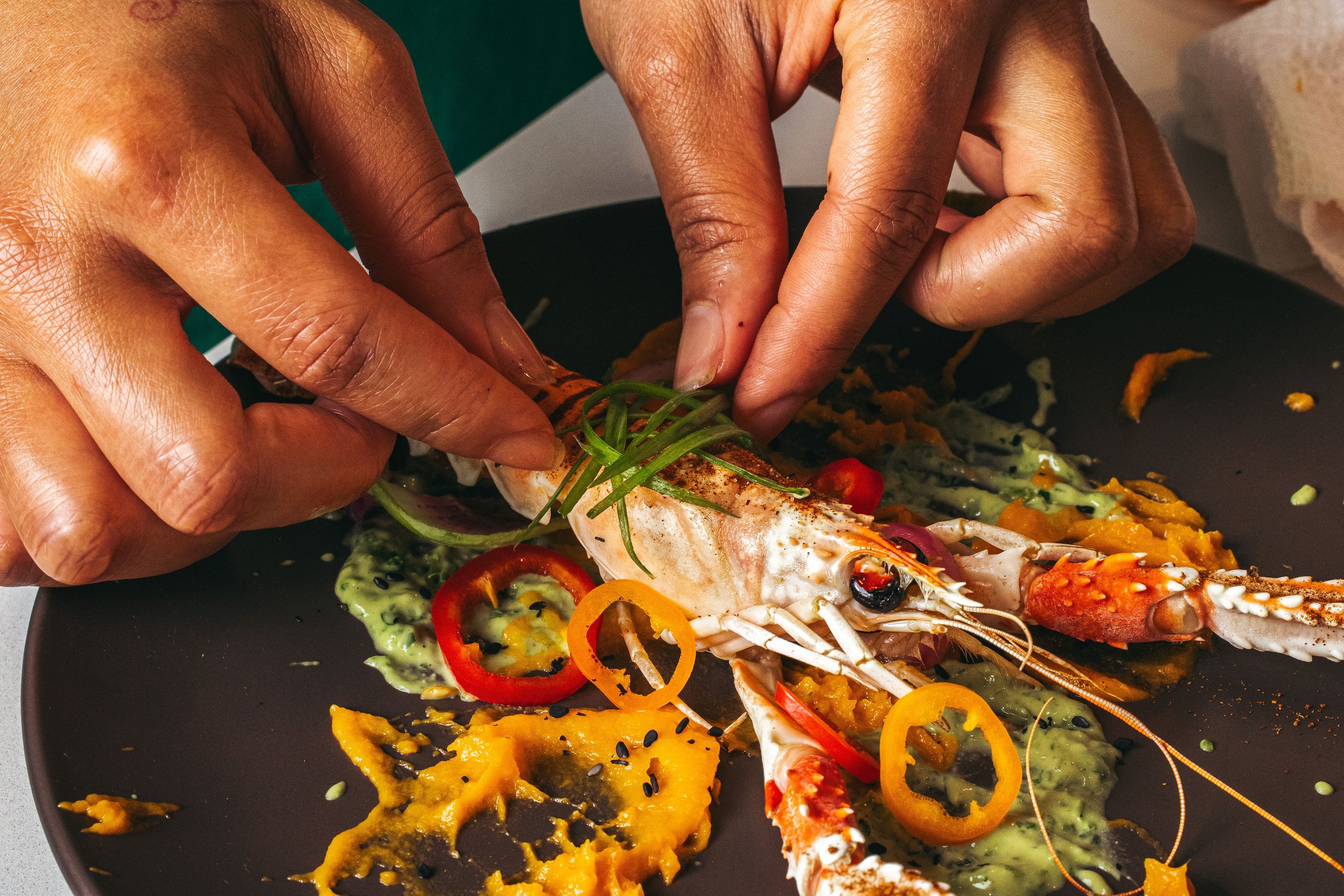 Hands plating a gourmet seafood dish with lobster, sliced red and yellow peppers, and green garnish on a black plate.