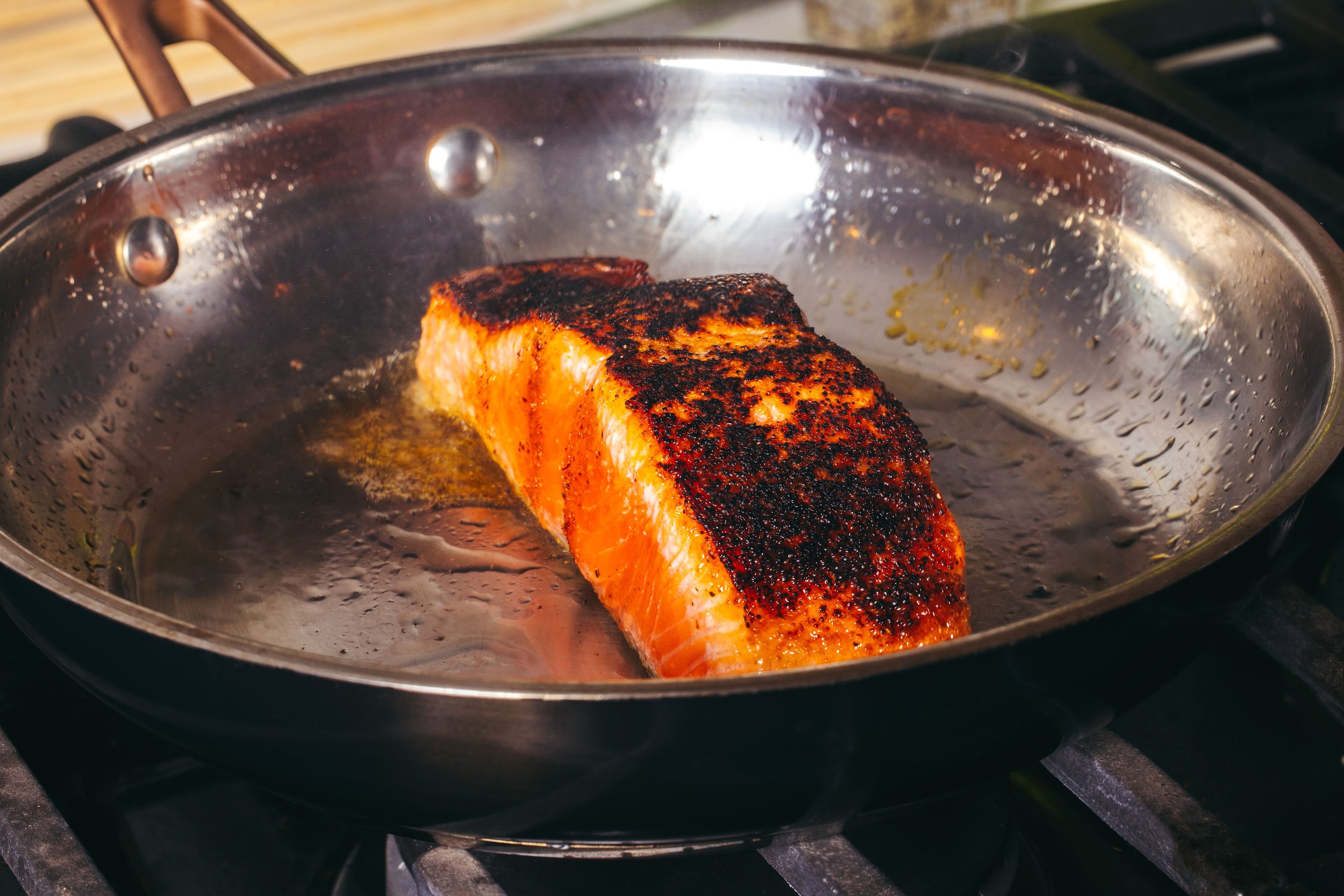 Salmon fillet cooking in a stainless steel frying pan with spices and oil