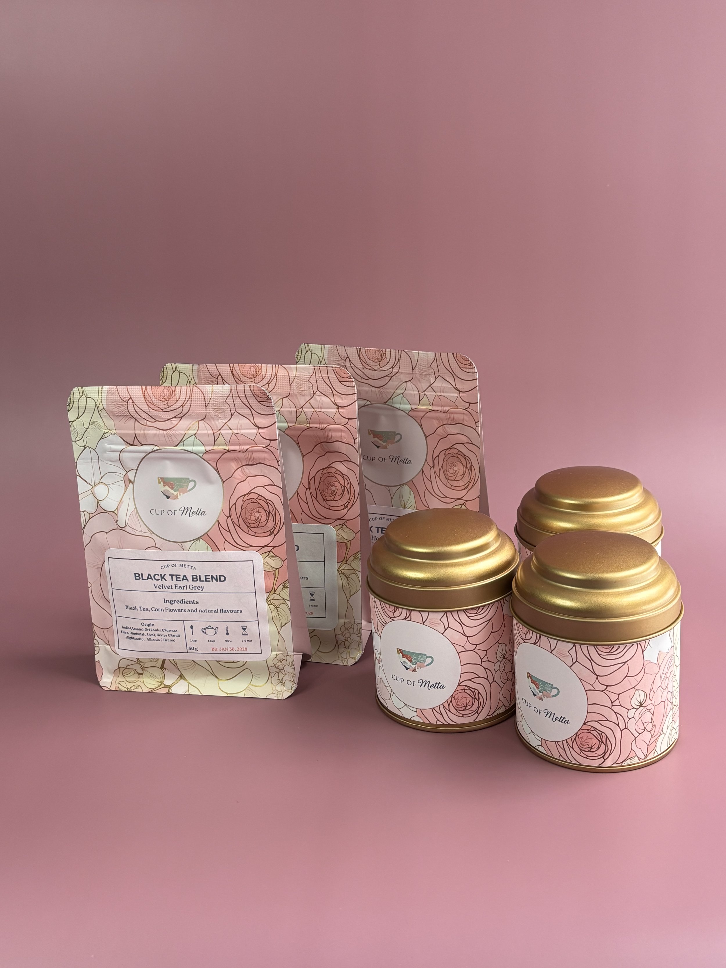 Three pink floral patterned tea tins with gold lids and three matching pink floral patterned tea packages, all branded 'Cup of Metta'