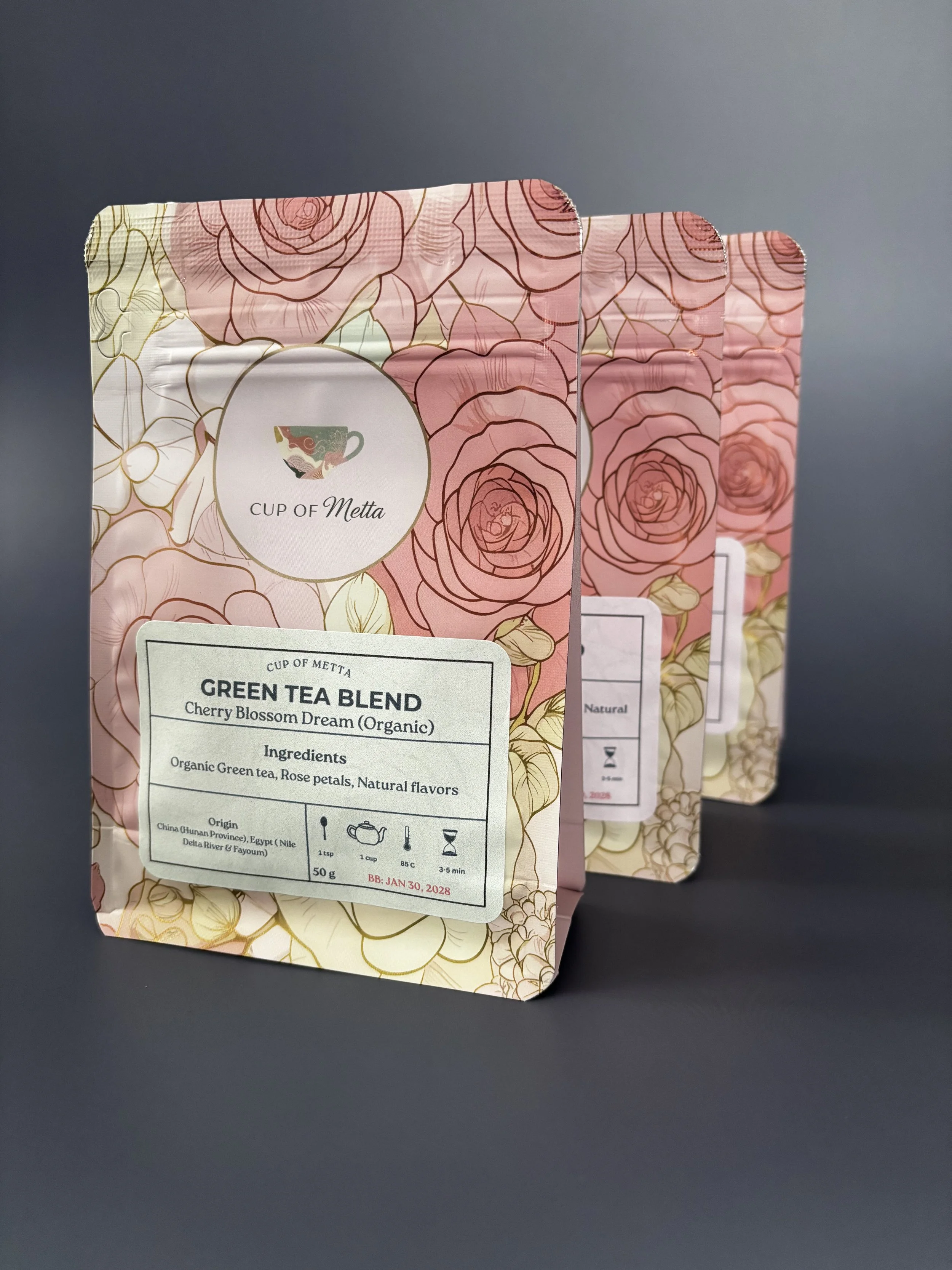 Three pink floral tea packets labeled 'Cup of Metta' with a rose and gold design, containing green tea blend with cherry blossom flavor.