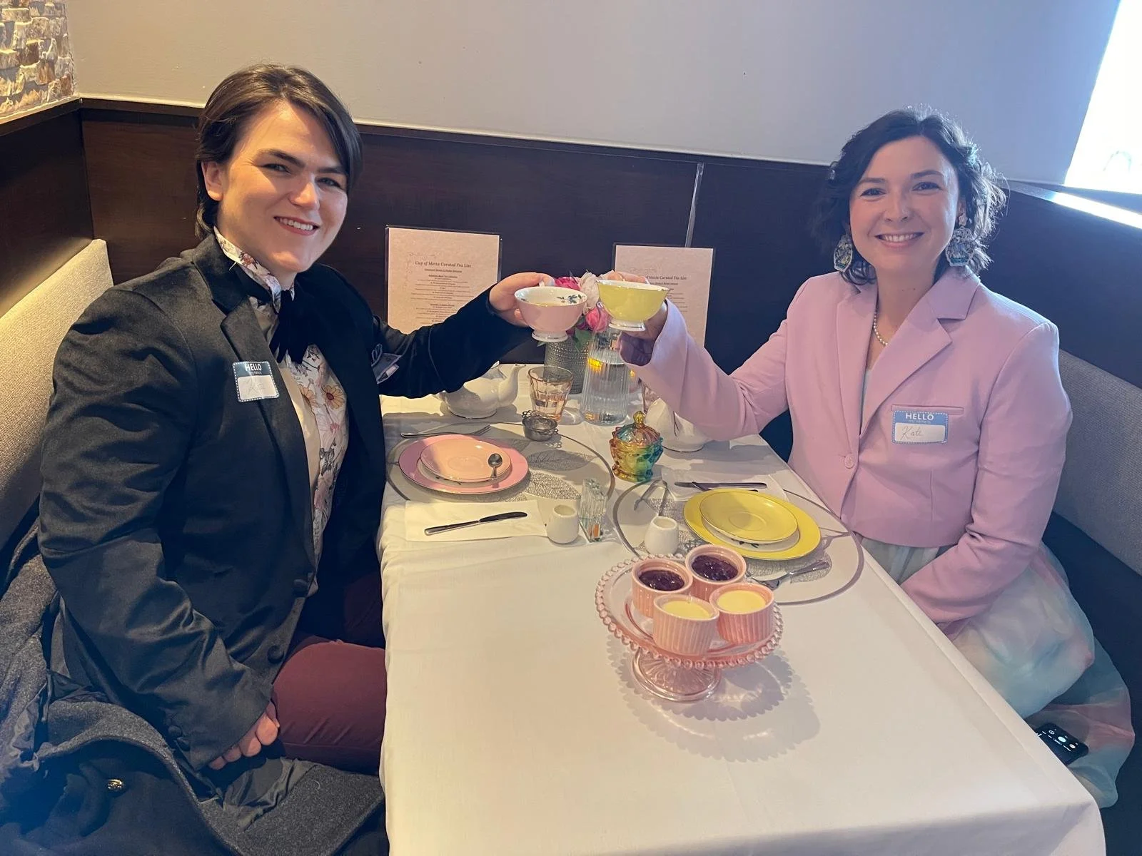 Two women celebrating at a tea party, clinking teacups, seated at a table with pastel-colored dishware and decorative items, smiling at the camera.