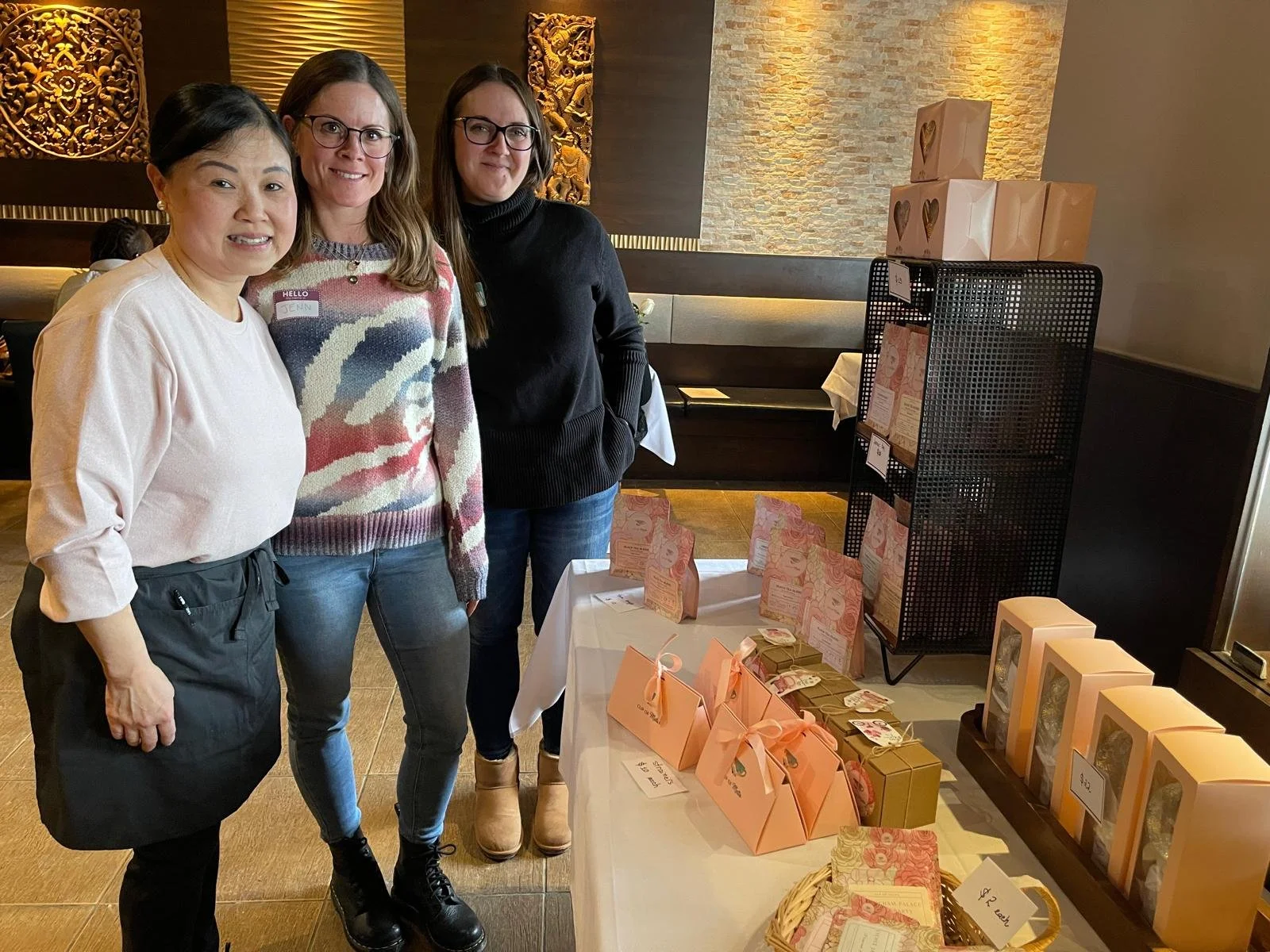 Three women standing together in a restaurant or cafe, smiling at the camera, with a table displaying various packaged gifts or products.