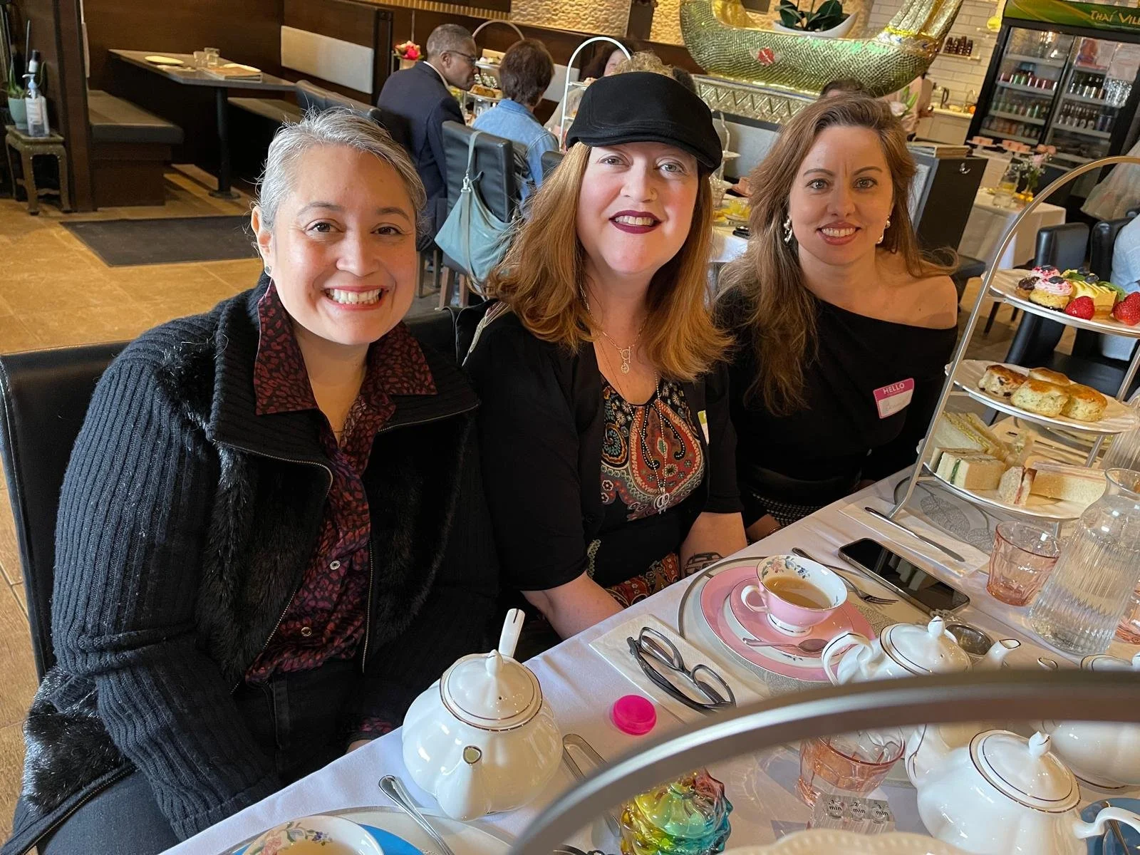 Three women sitting at a teacup and snack table in a restaurant, smiling at the camera, with a tiered tray of desserts and sandwiches in front of them.
