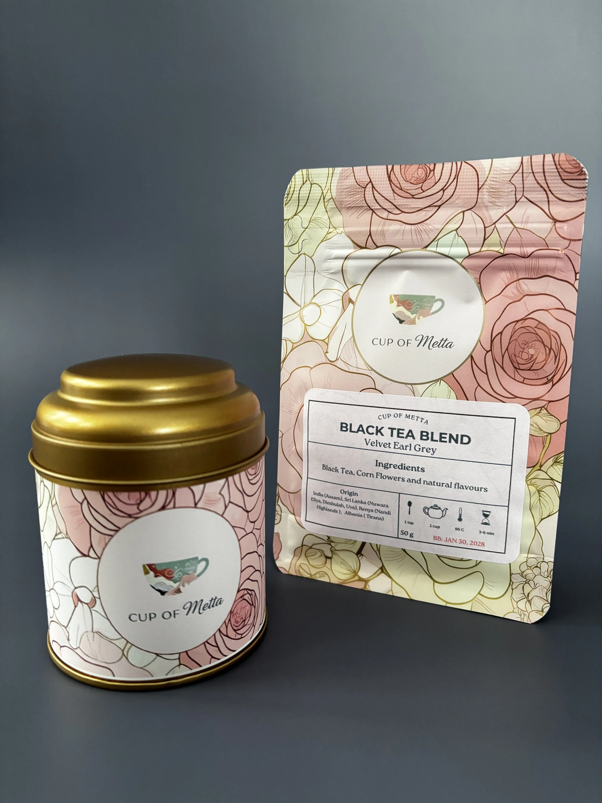 A pink floral patterned tea package and a matching tea tin with gold rim, both labeled 'CUP OF Metta'.