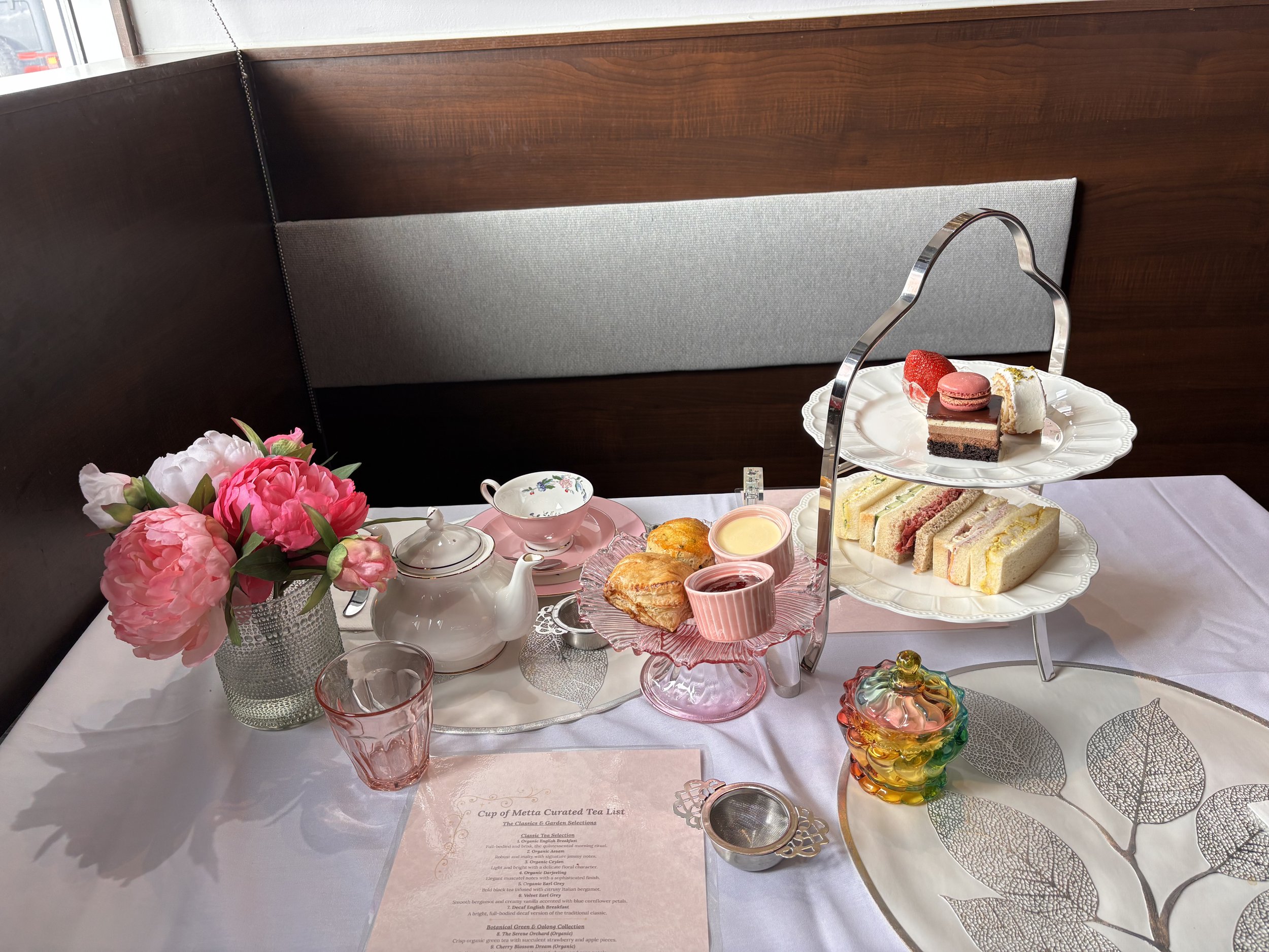 A pastel-colored illustration of a tea party setup with macarons and desserts on a three-tiered stand, a white teapot, and two cups of tea against a background of large pink and cream roses.
