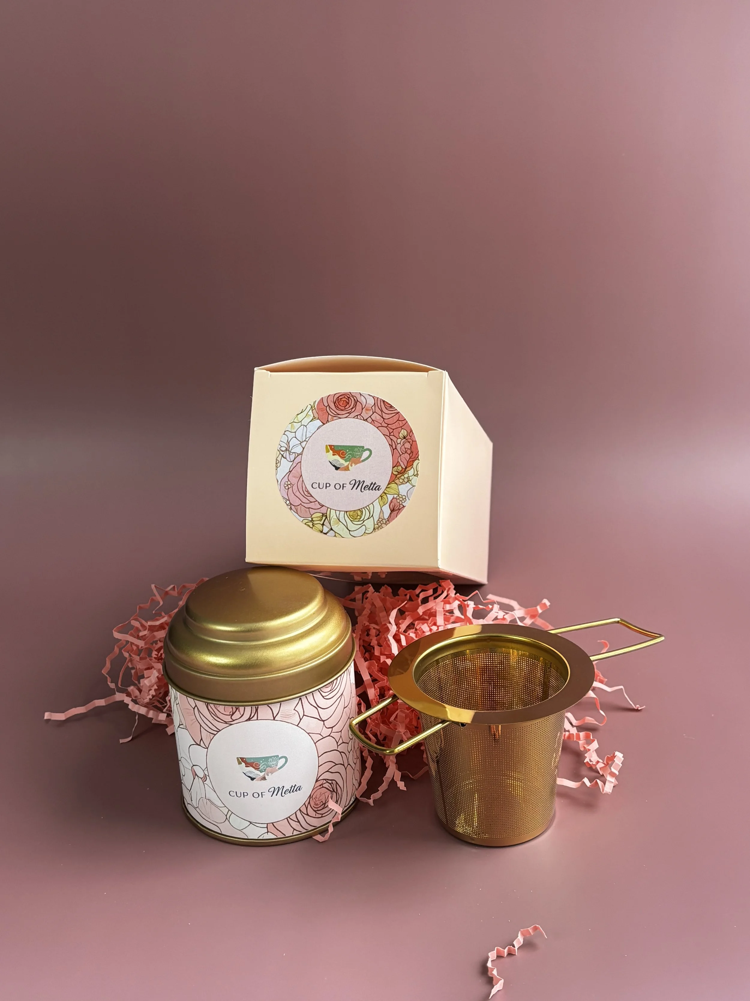 A tea infuser, a decorative tea tin, and a box with a floral design and a tea cup logo labeled 'Cup of Metta' on a pink background with pink shredded paper.