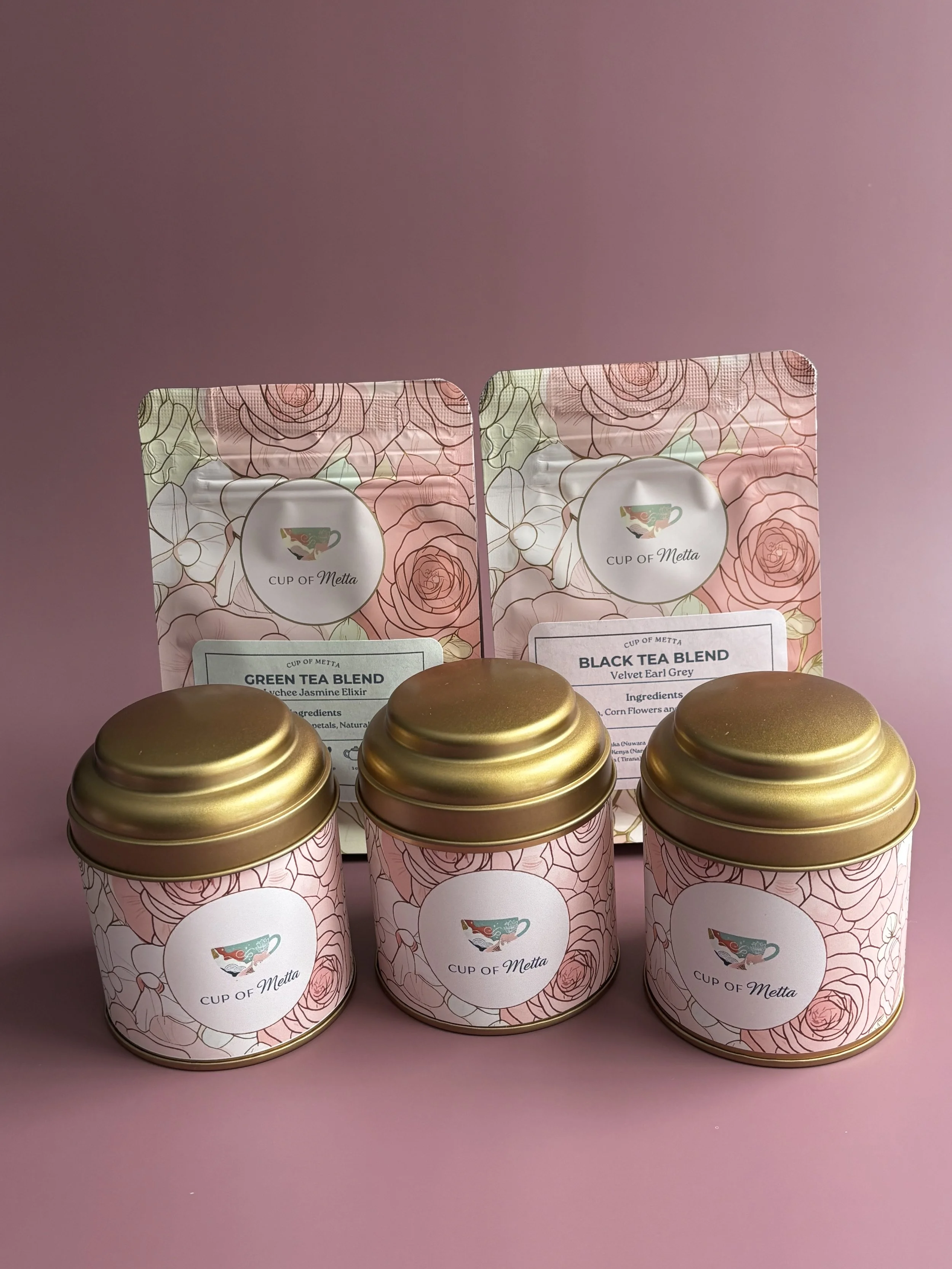 Three decorative tins with gold lids and pink floral patterns, labeled 'Cup of Metta,' and two pouches behind them labeled 'Green Tea Blend' and 'Black Tea Blend' with floral designs, set against a pink background.