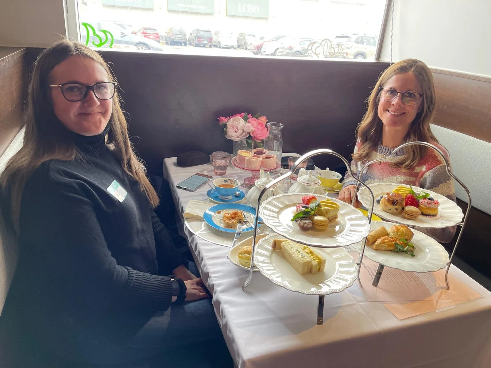 Two women sitting at a table in a restaurant with a two-tiered tray of assorted desserts, tea, and pink flowers, near a large window overlooking a parking lot.