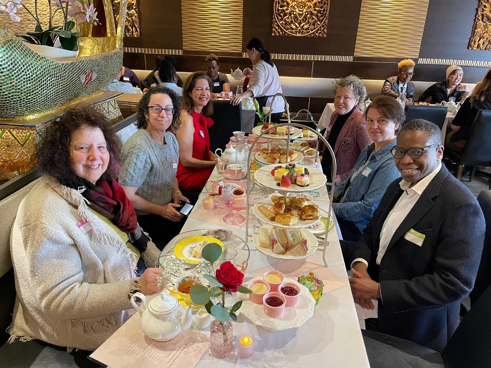 A group of seven diverse women and one man sitting at a long table in a restaurant, having a tea or brunch gathering. The table is set with various plates of sandwiches, pastries, cakes, and drinks, with some decorative flowers in the foreground.