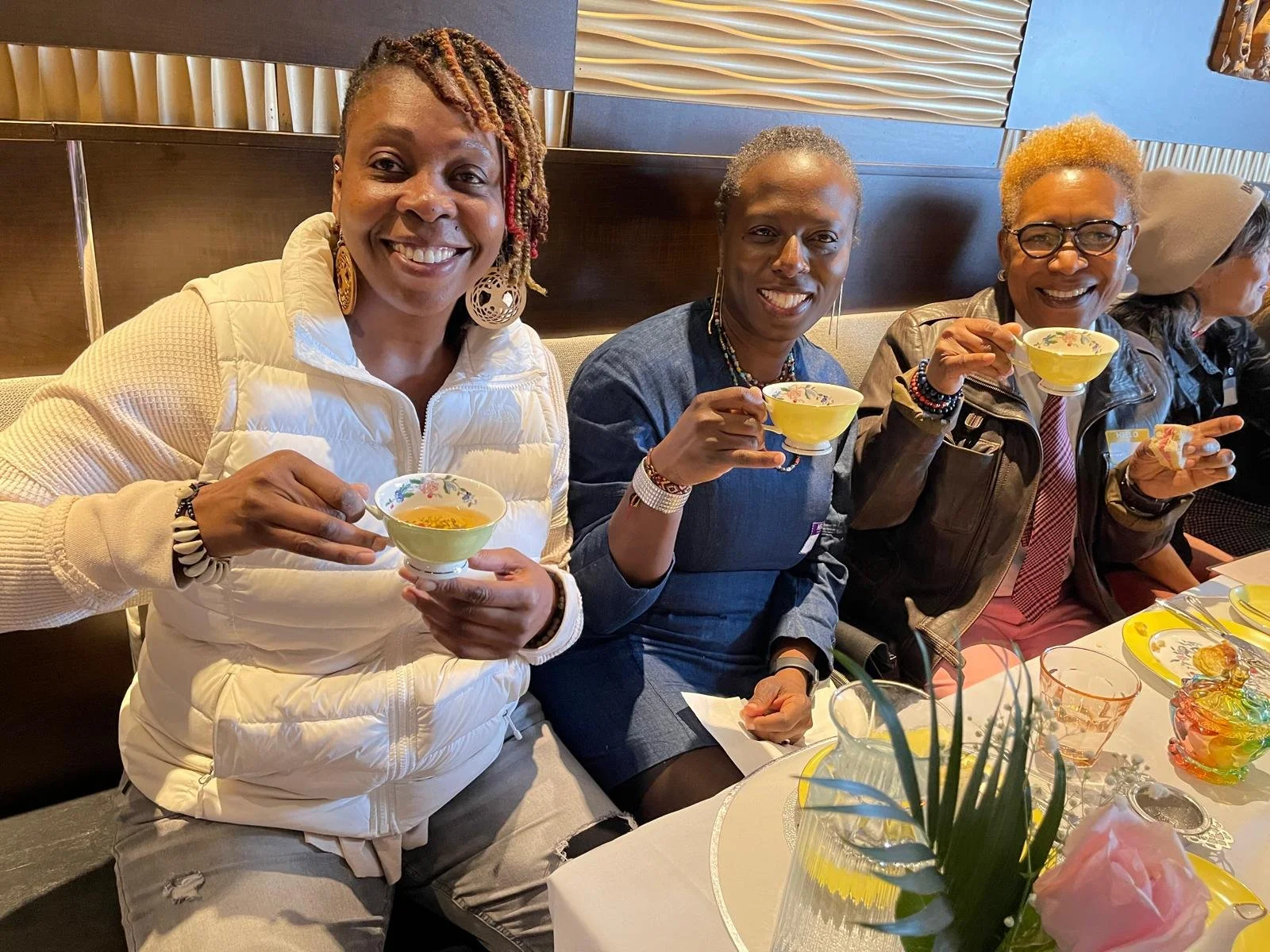 Four women sitting at a table, holding teacups, smiling, and enjoying a tea gathering in a restaurant.