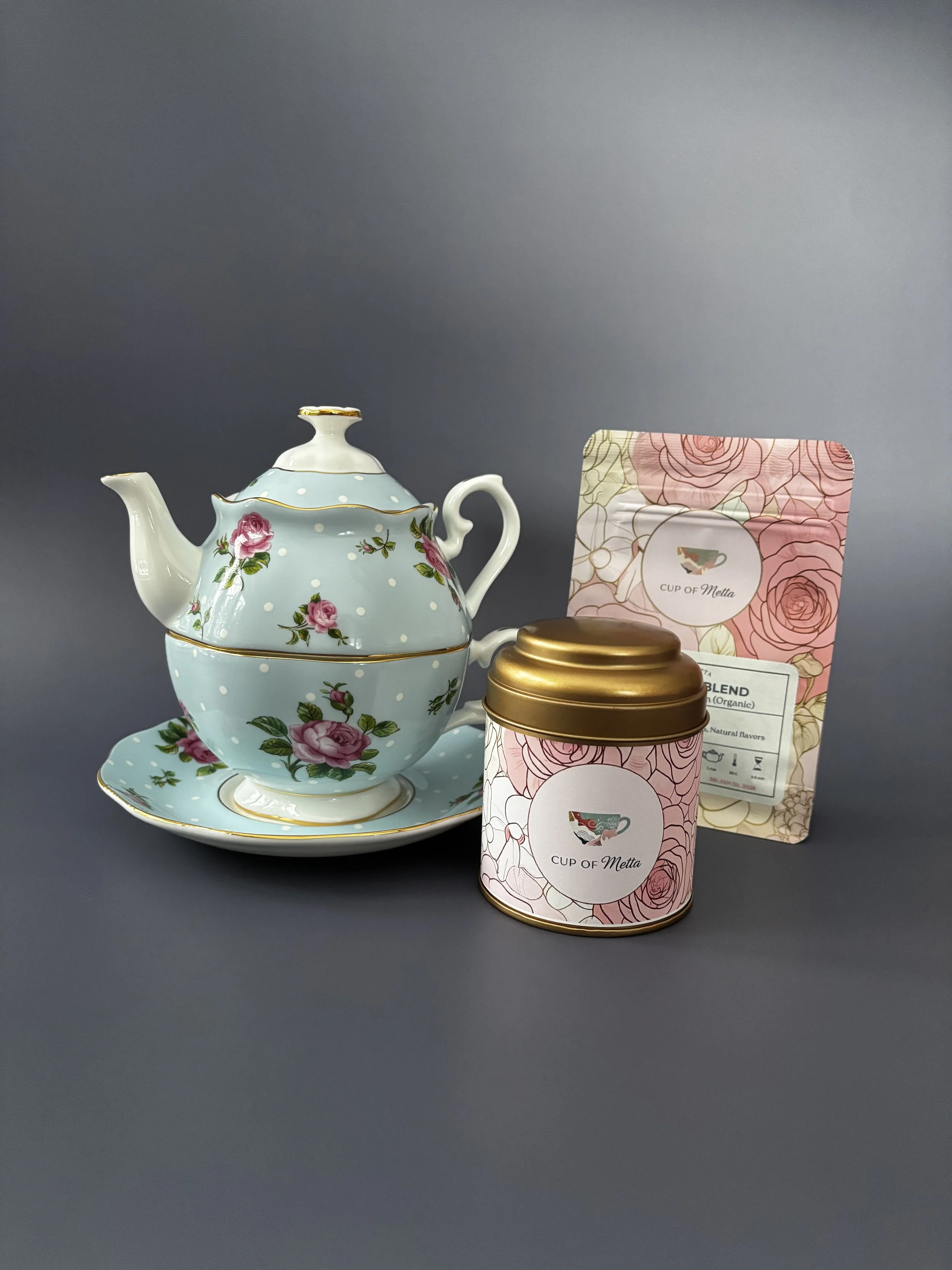 A teapot and matching cup and saucer with pink rose designs, along with a pink tin container and a packet of tea labeled "Cup of Metta," all placed on a dark grey background.