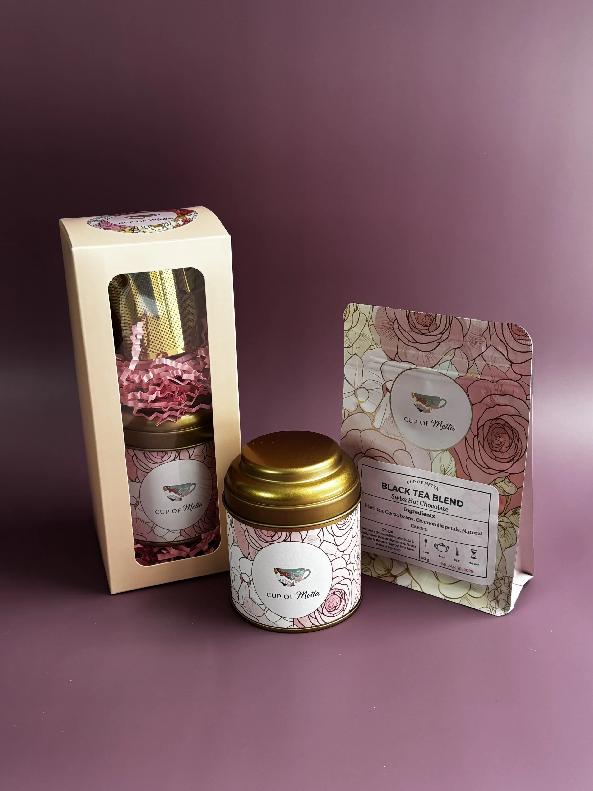 A boxed set of Swiss hot chocolate with a decorative floral design, including a jar, a packet labeled 'Black Tea Blend,' and a small gift box, all arranged on a pink background.