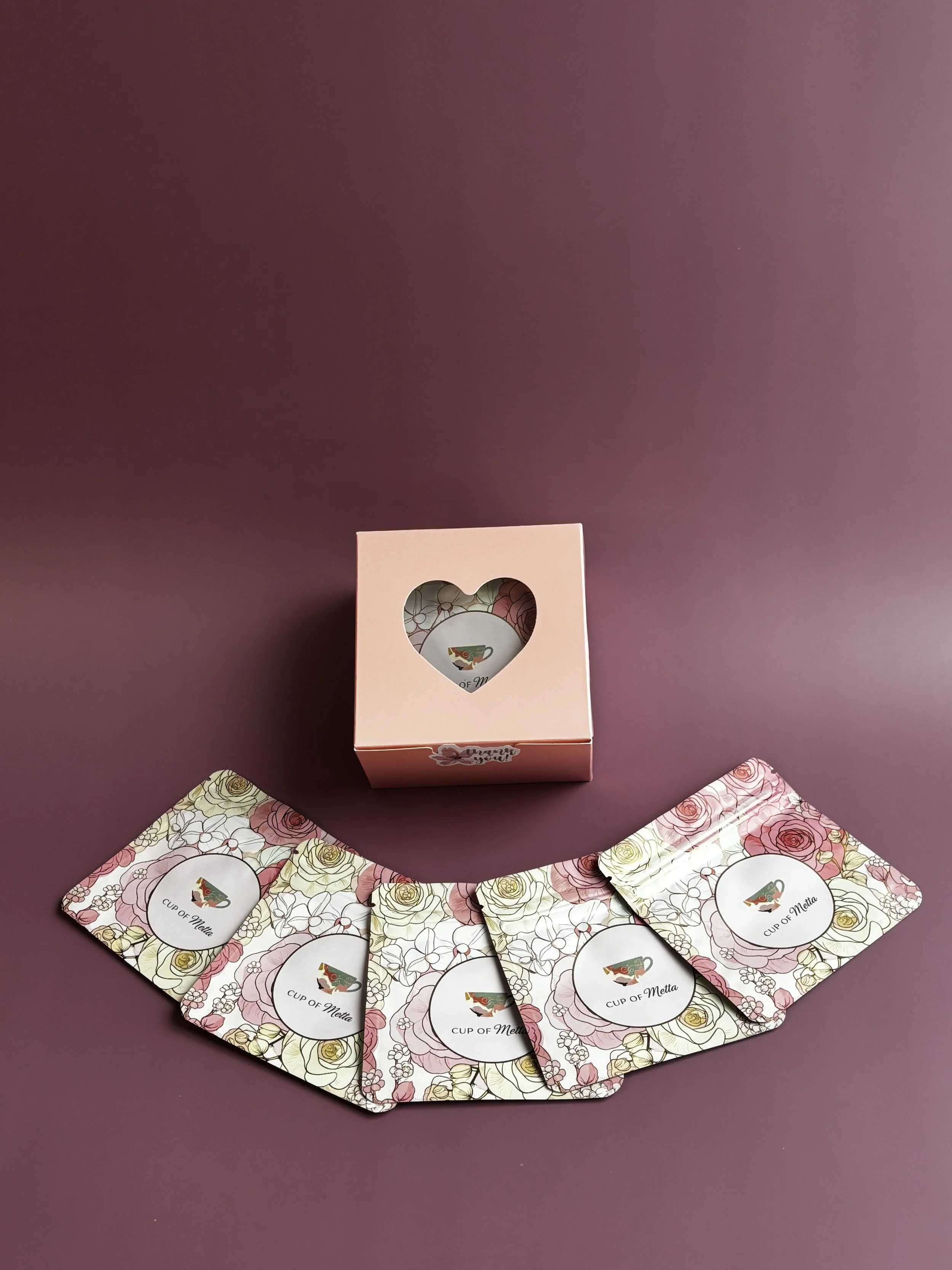 A pink box with a heart-shaped cutout window shows it is a tea set gift box. Five floral-patterned cards with the label 'Cup of Meta' are arranged in a semi-circle on a purple surface.