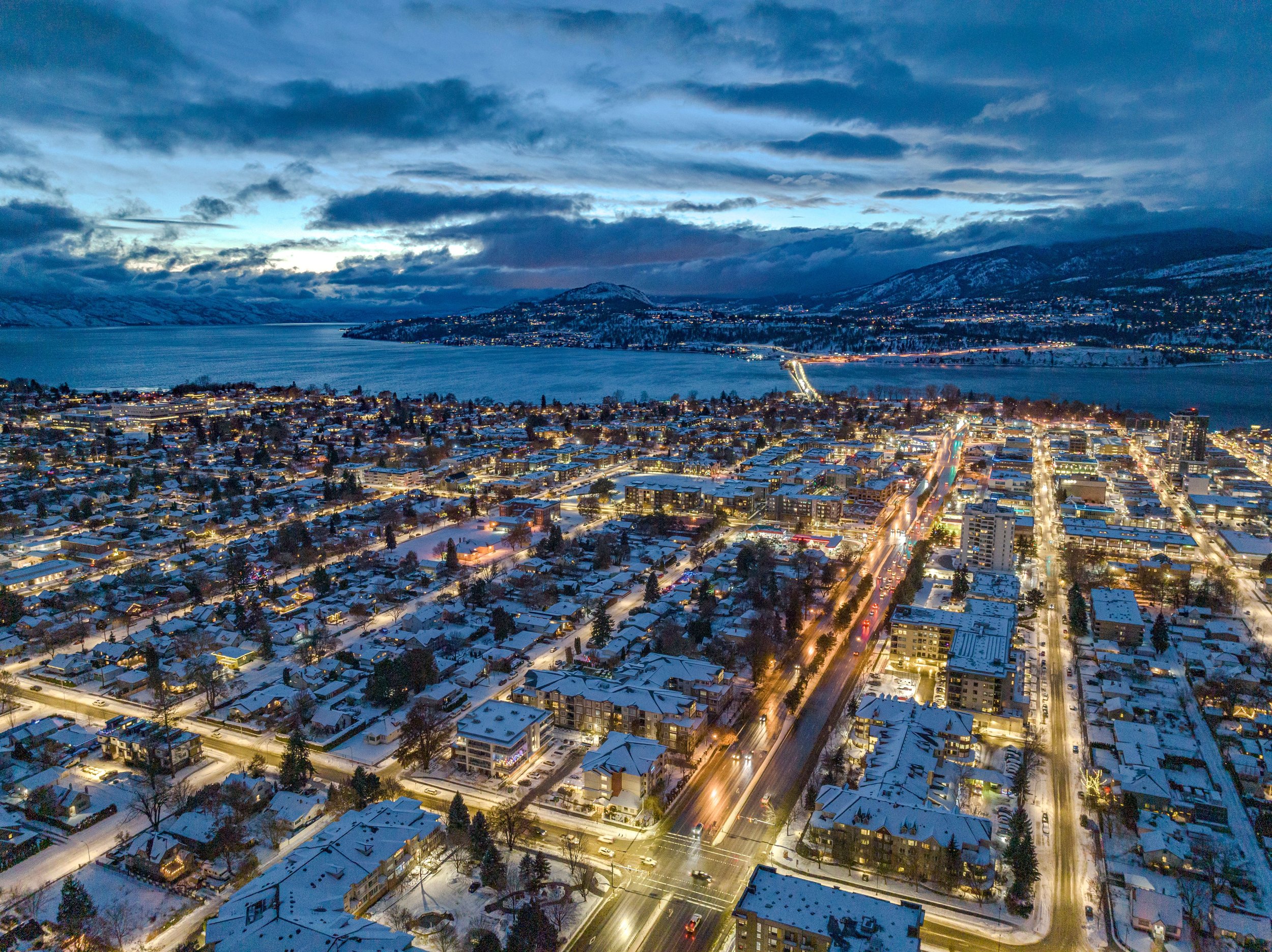 An aerial view of a snow-covered downtown Kelowna during twilight with a large body of water and mountains in the background, city streets illuminated by streetlights.