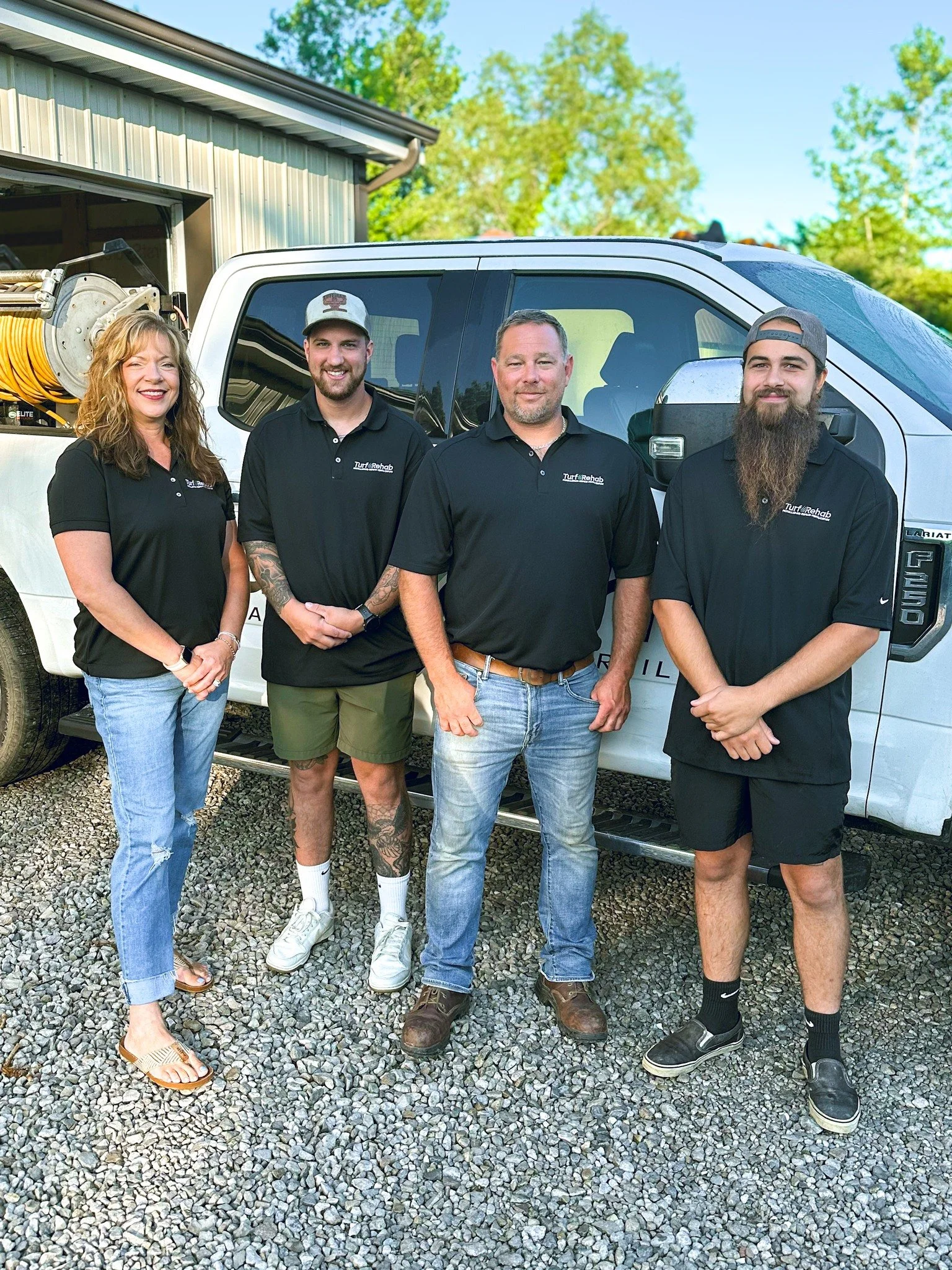 Four people standing in front of a white utility truck on a gravel driveway, smiling at the camera. They are wearing black polo shirts with a company logo, with a woman on the left and three men with different hairstyles and facial hair.