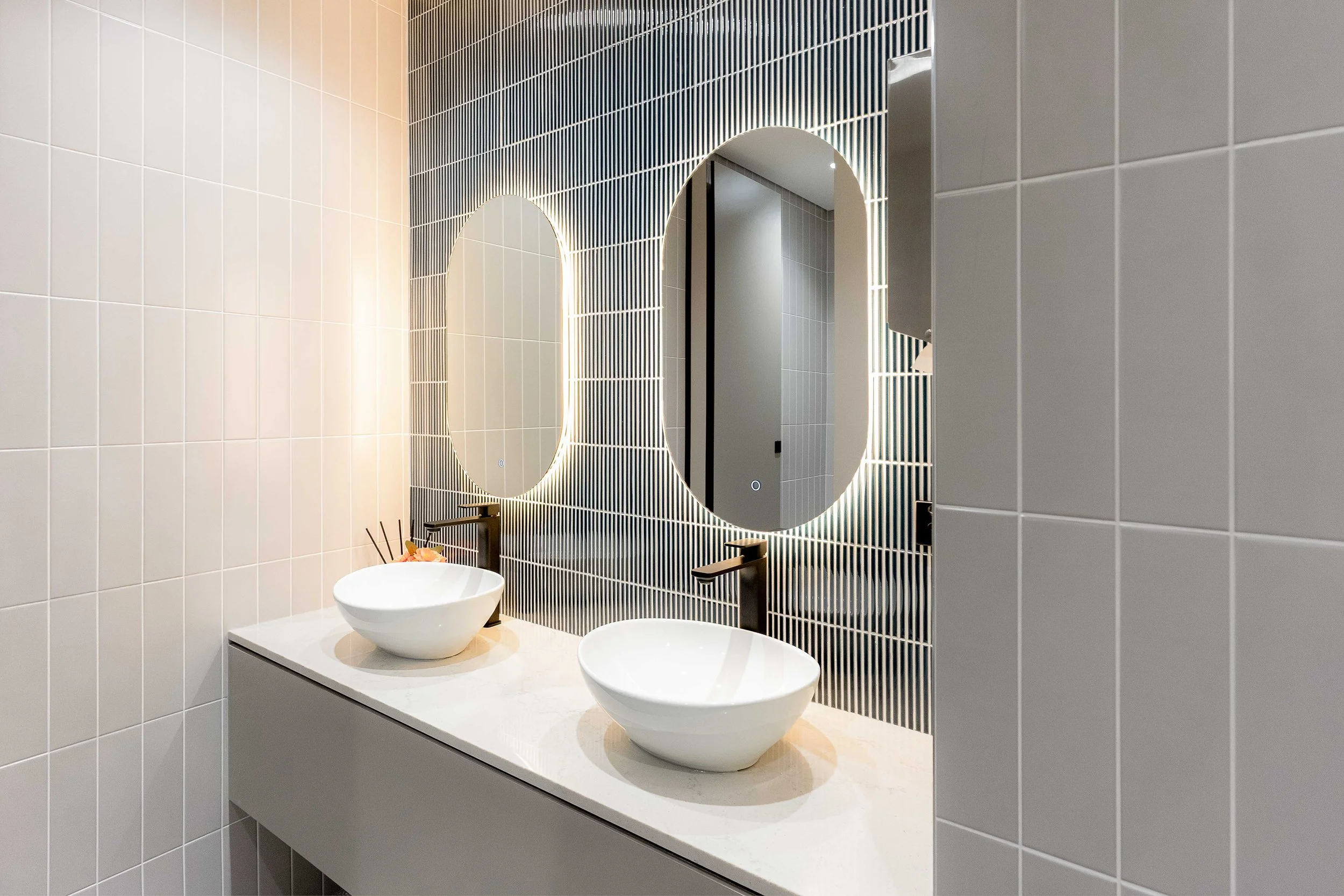 Modern bathroom with two white vessel sinks on a white countertop, a mirror with LED lighting, and tiled walls in white and dark shades.