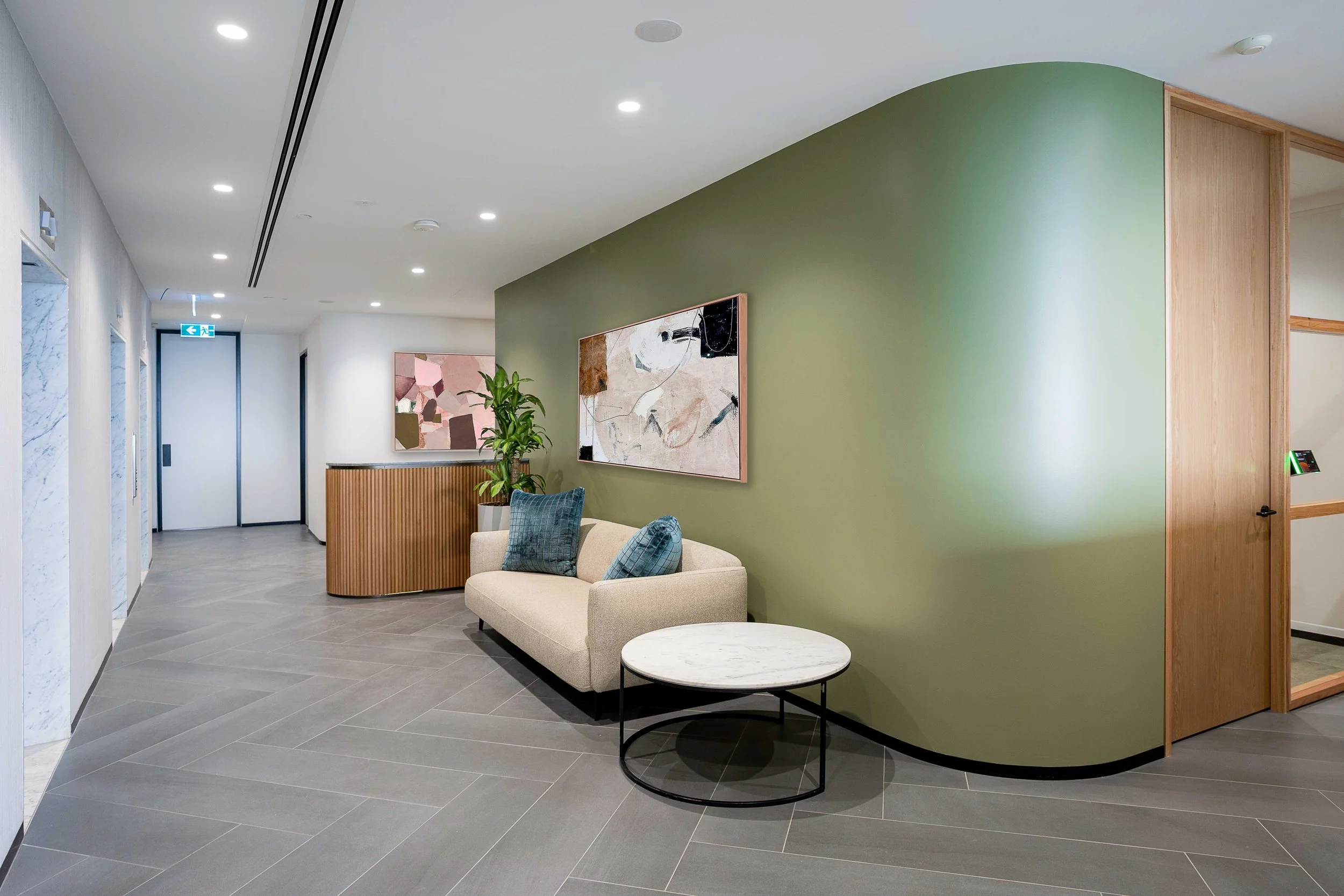 Interior of a modern lobby with a green curved accent wall, a beige sofa with blue pillows, a small round marble table, and artwork on the wall. The floor is tiled in gray, and the ceiling has recessed lighting and an emergency exit sign in the background.