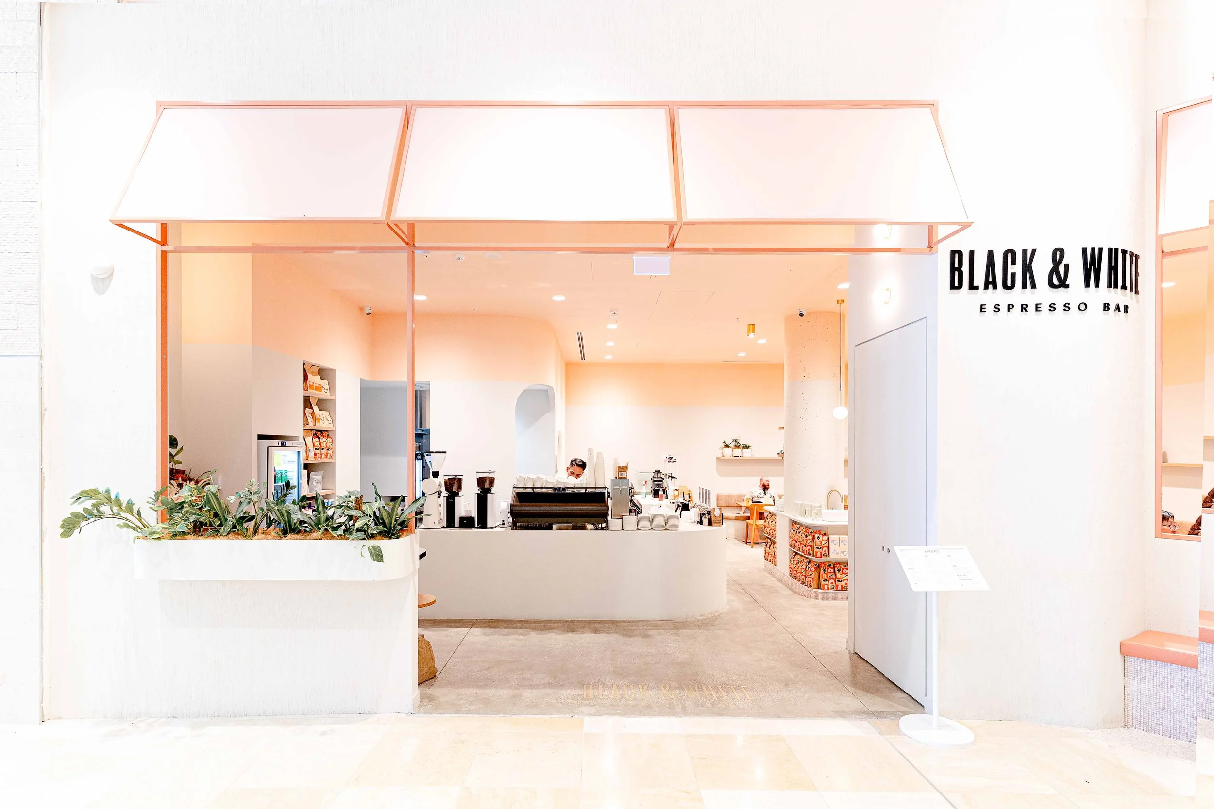 Entrance to Black & White Espresso Bar with a white and pink color scheme, visible inside are coffee machines, cups, and a staff member working.
