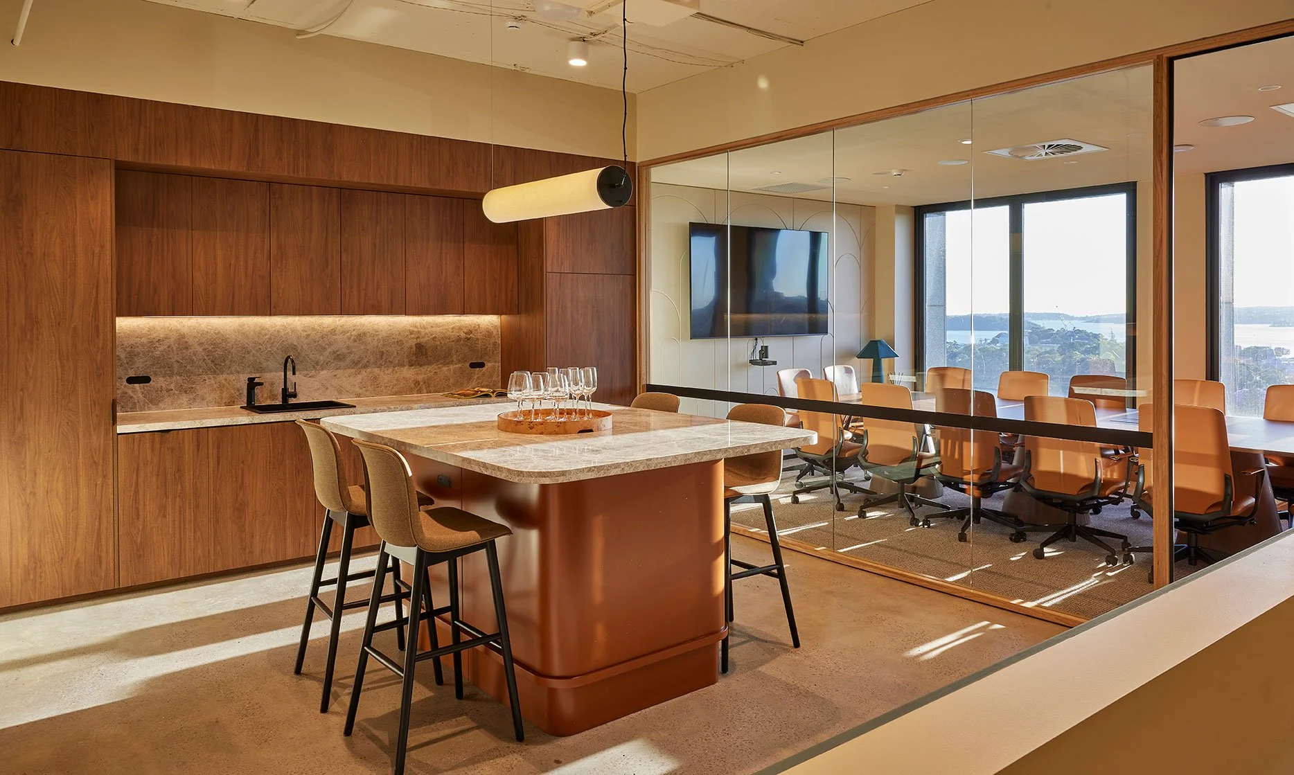 Modern conference room with large windows, multiple chairs around a long table, and a wall-mounted TV. A kitchenette with a marble countertop and cabinets is visible in the foreground, with a tray of glasses on the counter. The room is well-lit with natural light.