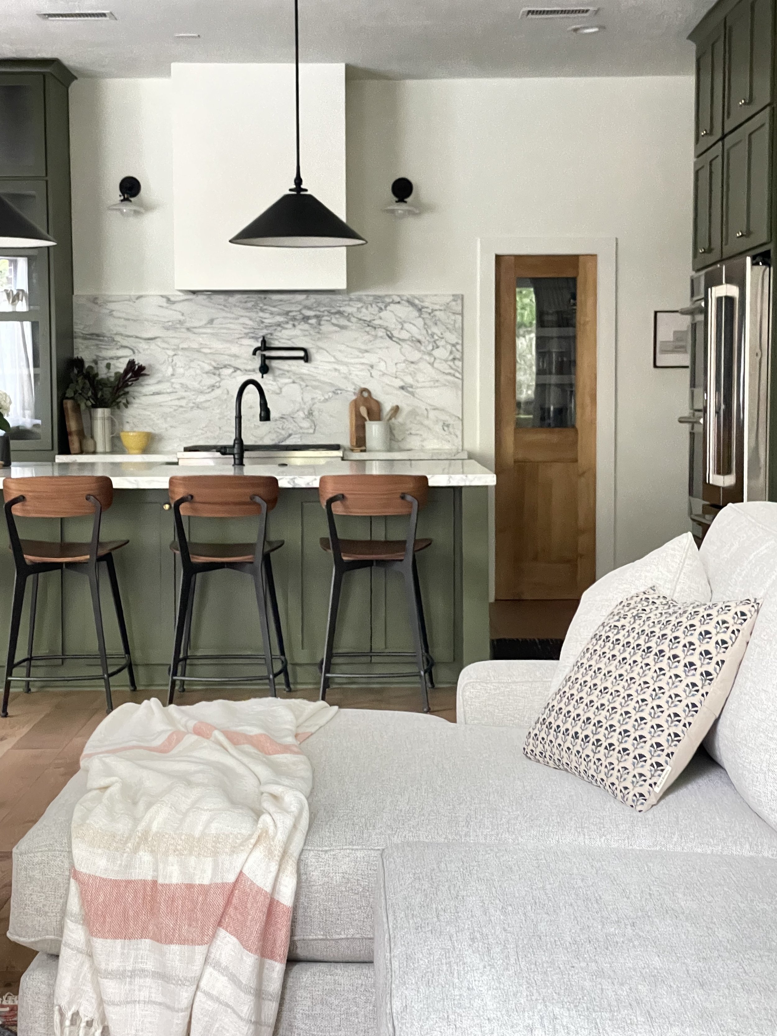 Living room with a white couch, decorative pillows, and a throw blanket, overlooking a green kitchen island and bar stools with a marble backsplash and wooden accents.