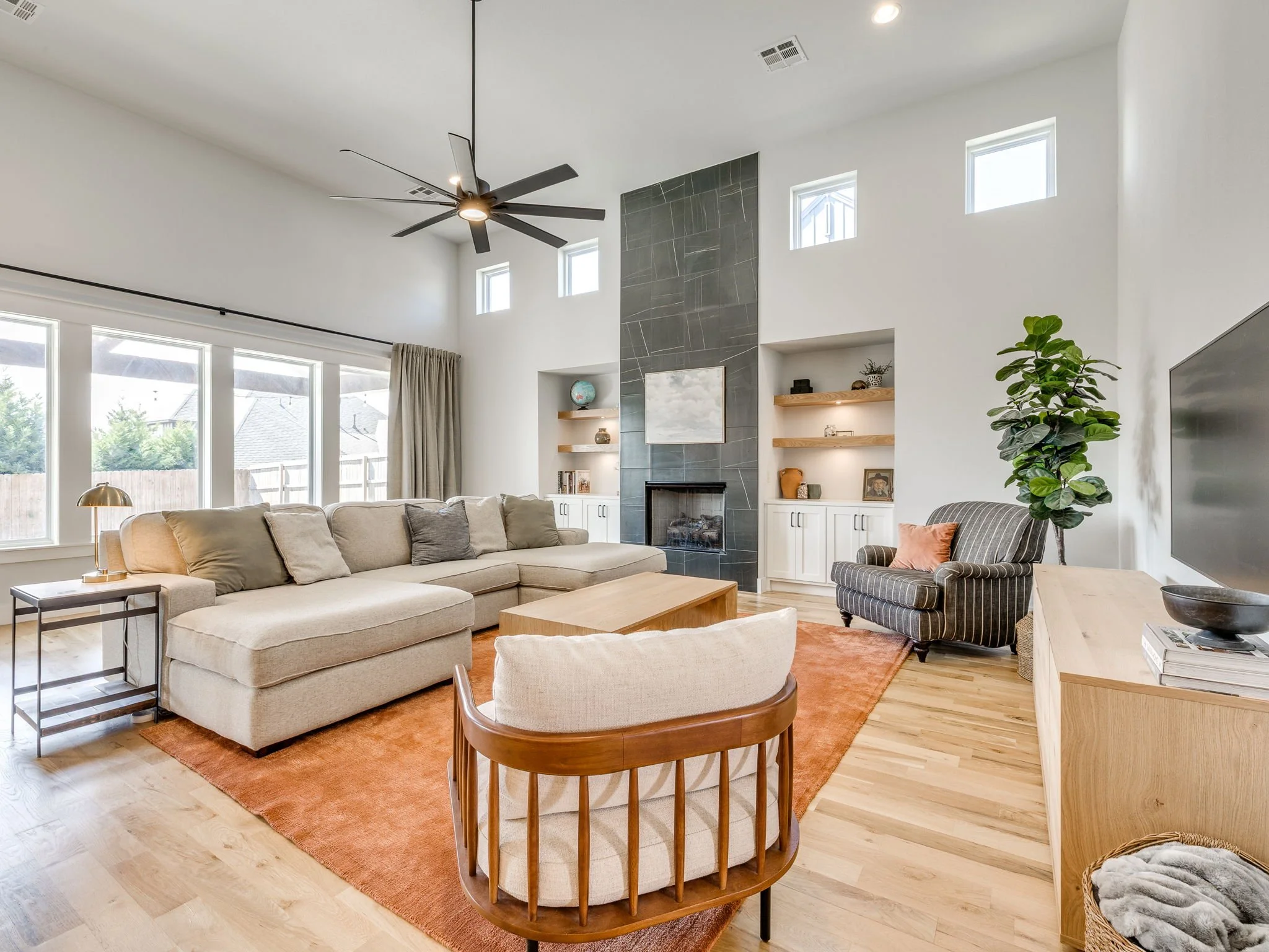 Living room with beige sectional sofa, orange area rug, black ceiling fan, tall windows, built-in shelves with decor, and a potted plant.
