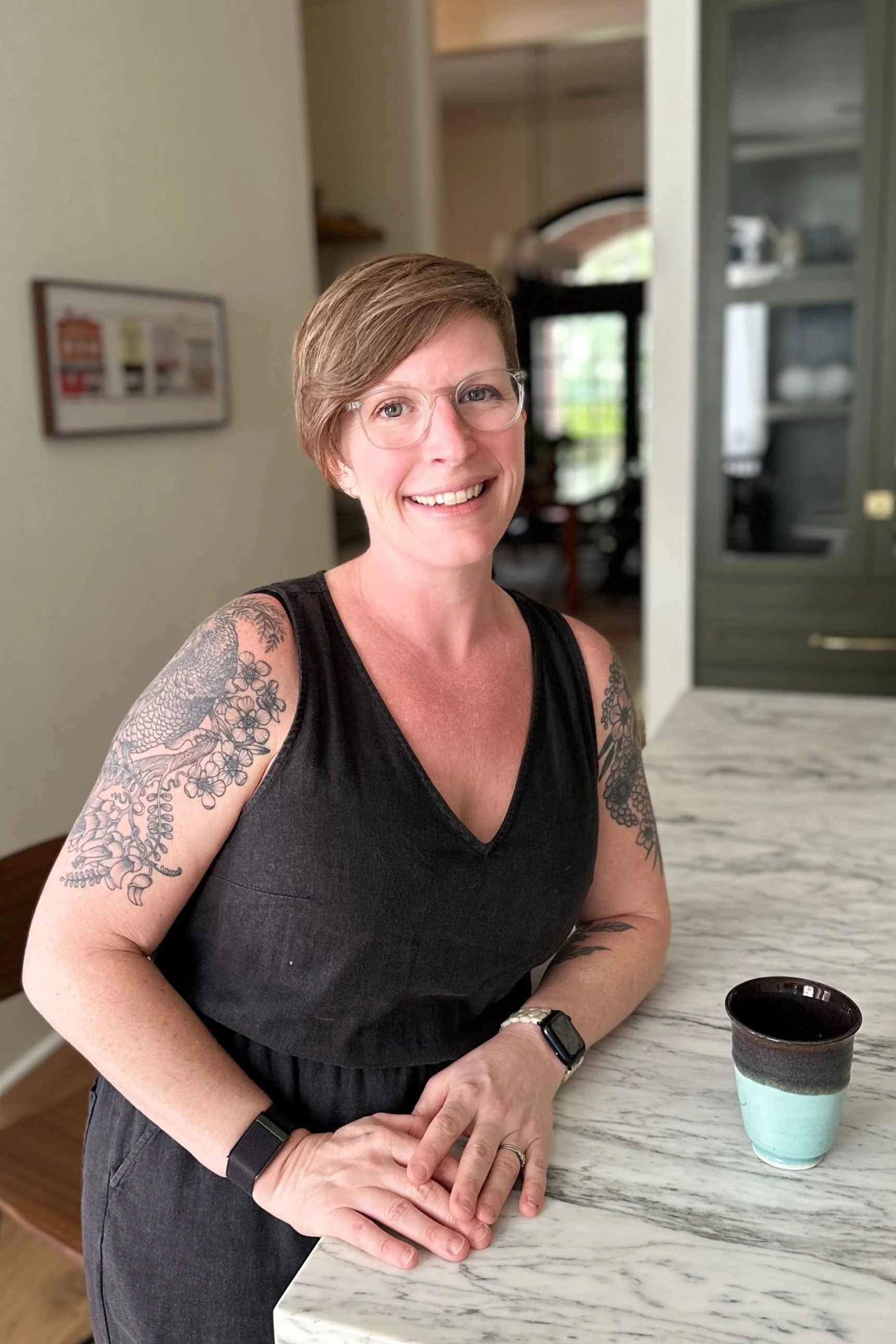A smiling woman with short brown hair, glasses, and tattooed arms standing at a kitchen counter with a cup of coffee.