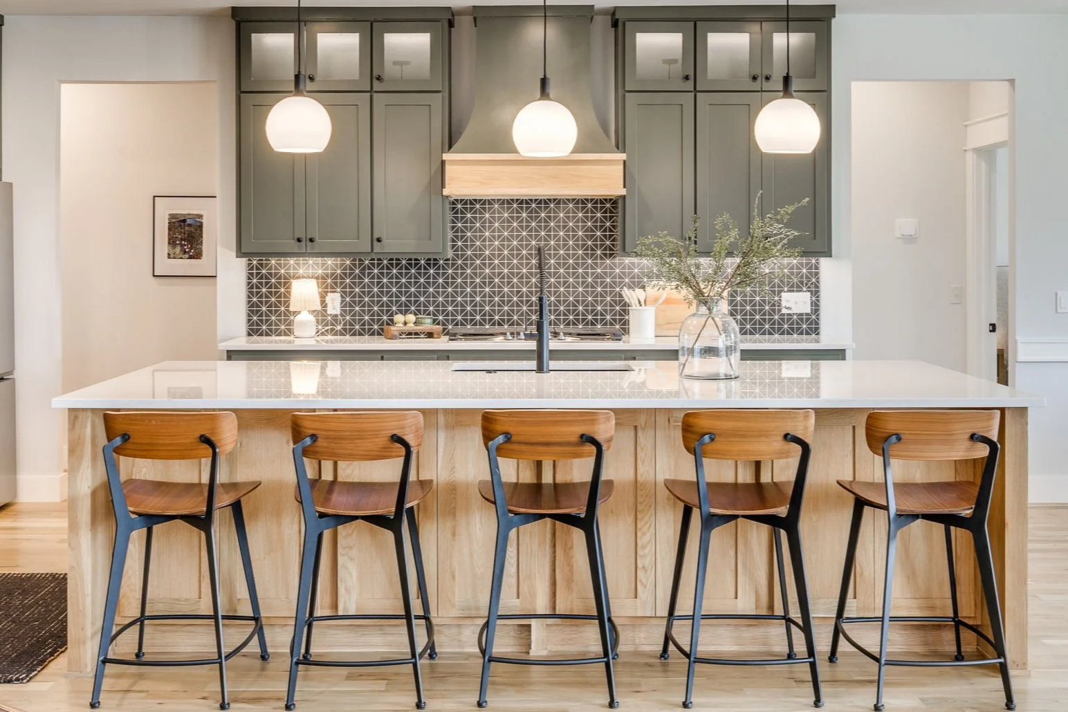 Modern kitchen with green cabinetry, black and white geometric backsplash, white countertop island with five wooden chairs, pendant lighting, and decorative elements.