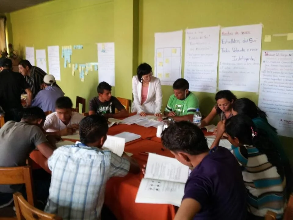 Group of students and a teacher gathered around a table with books, in a classroom with posters and charts on the wall.