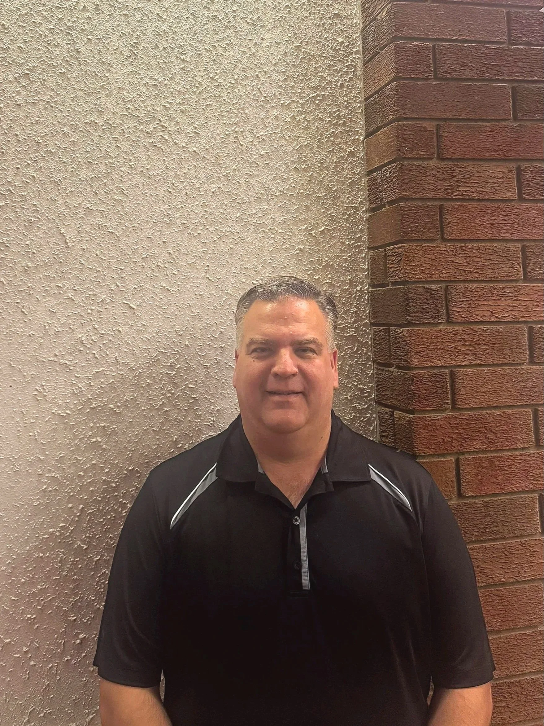 A man with short gray hair wearing a black polo shirt posing in front of textured beige wall and red brick wall.