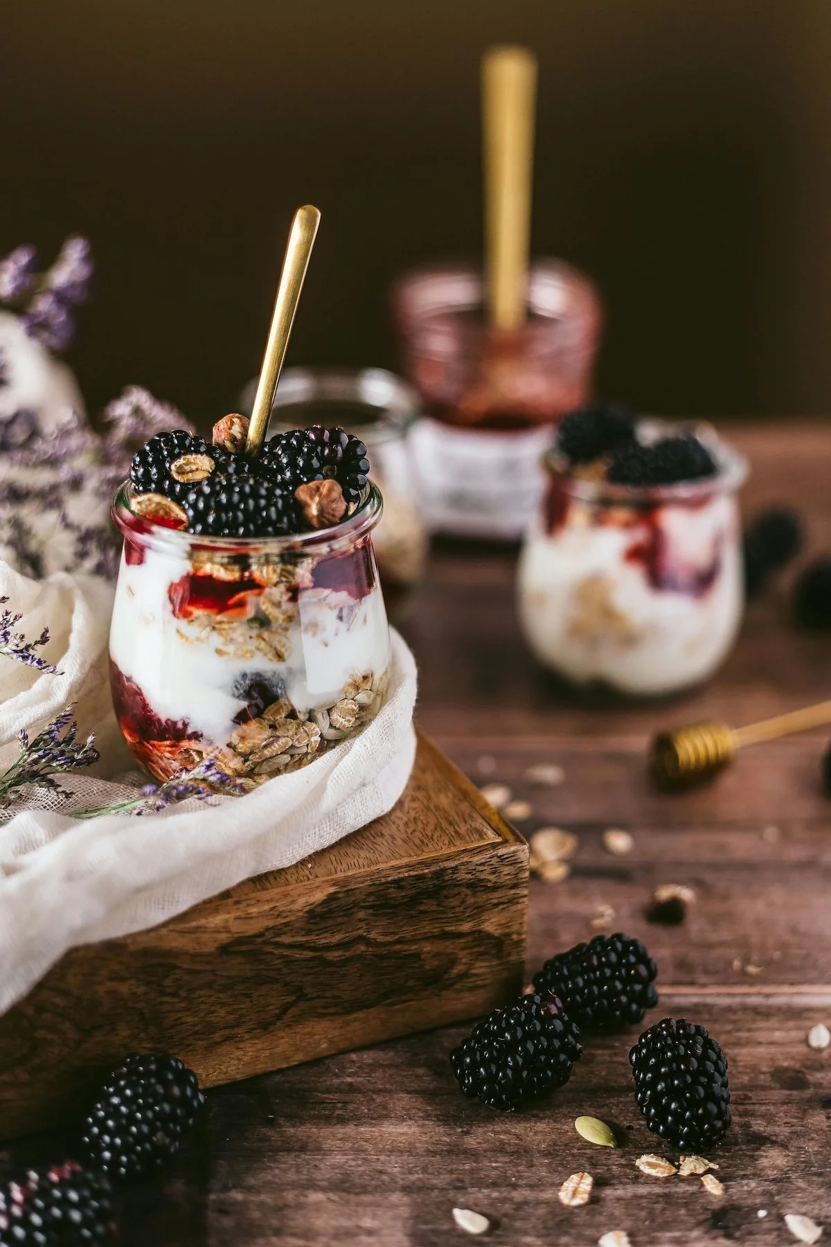 A glass jar filled with layered yogurt, blackberries, oats, and jam, topped with blackberries and nuts, placed on a wooden surface with scattered blackberries and oats nearby, and a small honey dipper in the background.