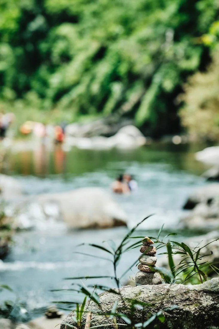 Stacked rocks in the foreground near a river with people swimming and relaxing in the background.