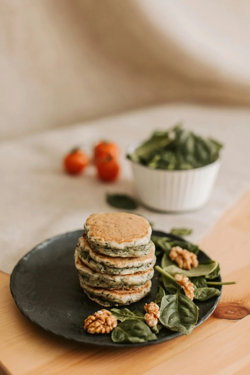 A black plate with a stack of four spinach and cheese savory pancakes garnished with leafy greens and walnuts. In the background, a white bowl filled with fresh spinach and a few cherry tomatoes on a light-colored surface.