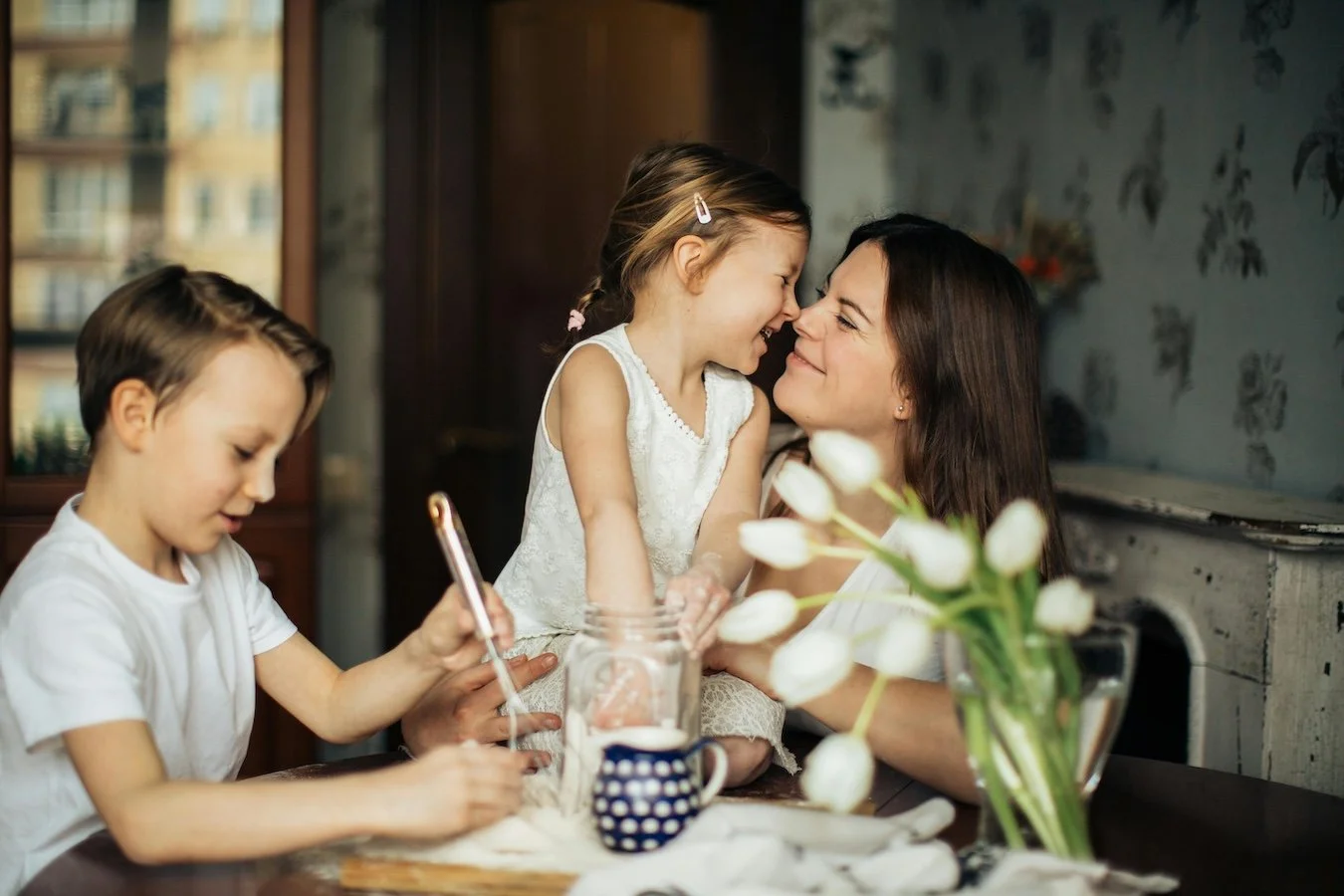 A mother and her young daughter are smiling and touching foreheads while sitting at a table with her son who is playing with a phone. The table has a vase with white tulips and a mug, and there is a window and framed picture in the background.