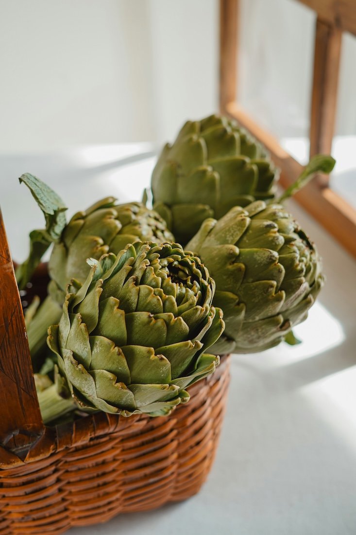 healthy eating with nutritionist Bjorn Paech represents Four fresh artichokes in a wicker basket on a white surface near a window.