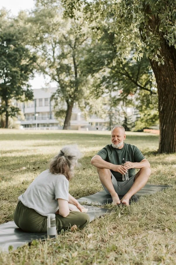 An elderly woman and a man practicing yoga or meditation outdoors on a grassy area under a tree, with a water bottle nearby and buildings in the background.