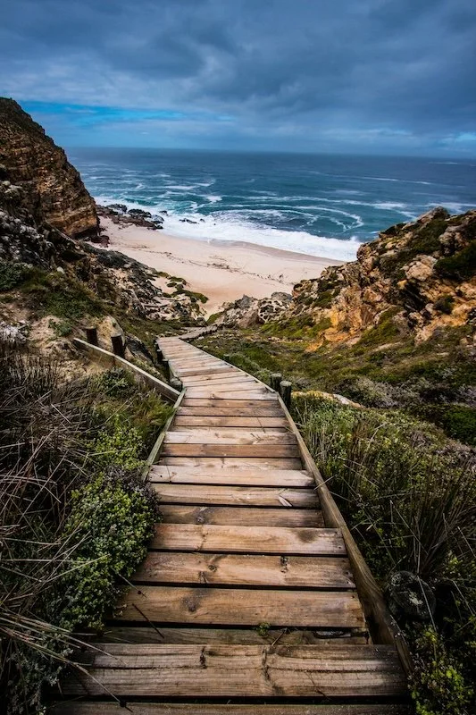 Yeppoon naturopath, Bjorn Paech shows a Wooden pathway leading down to a sandy beach with ocean waves under a cloudy sky.
