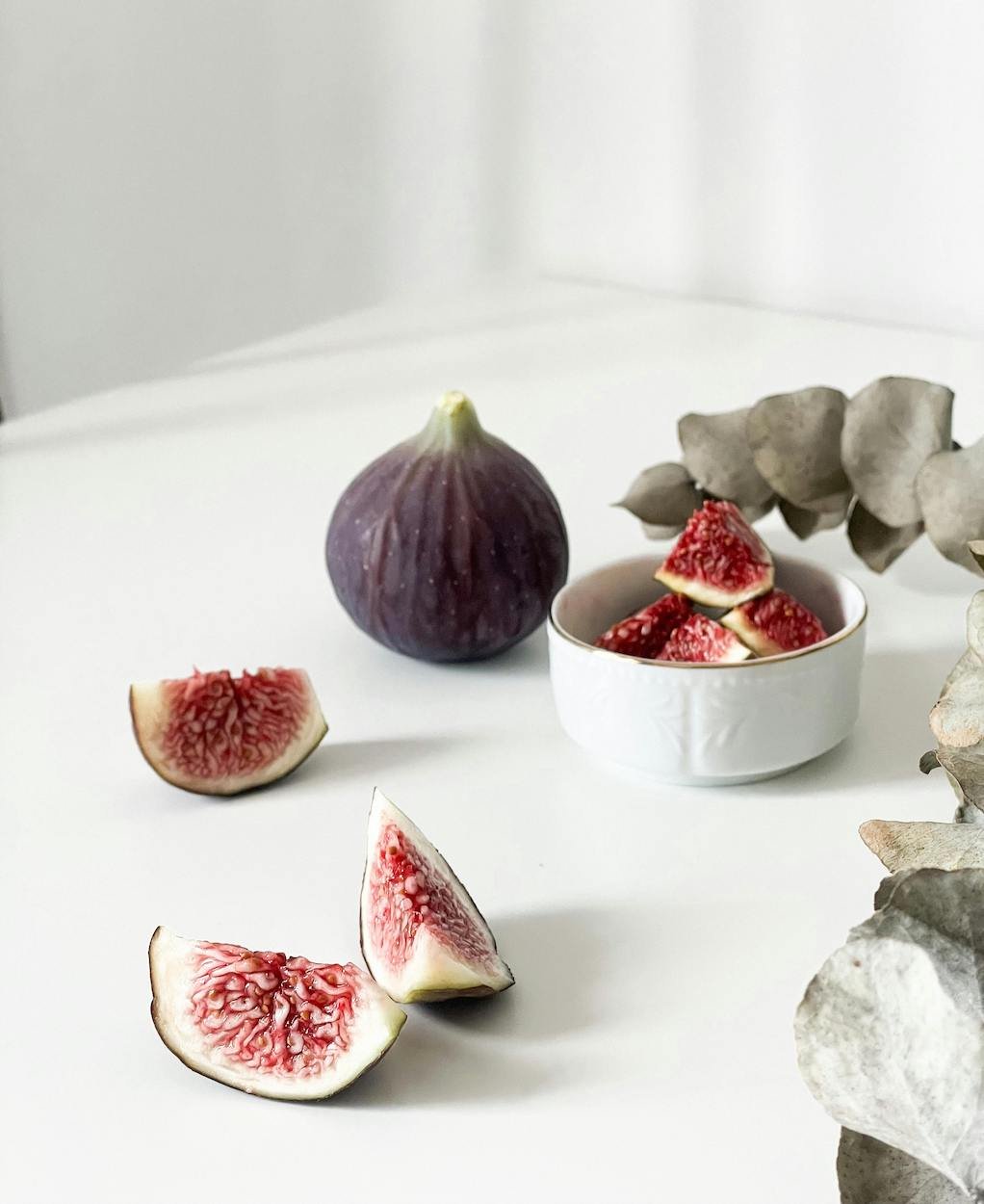 Nutritionist for women yeppoon shows a bowl of Fresh purple figs, some sliced and some whole, arranged on a white surface with a bowl of sliced figs and gray-green dried leaves.