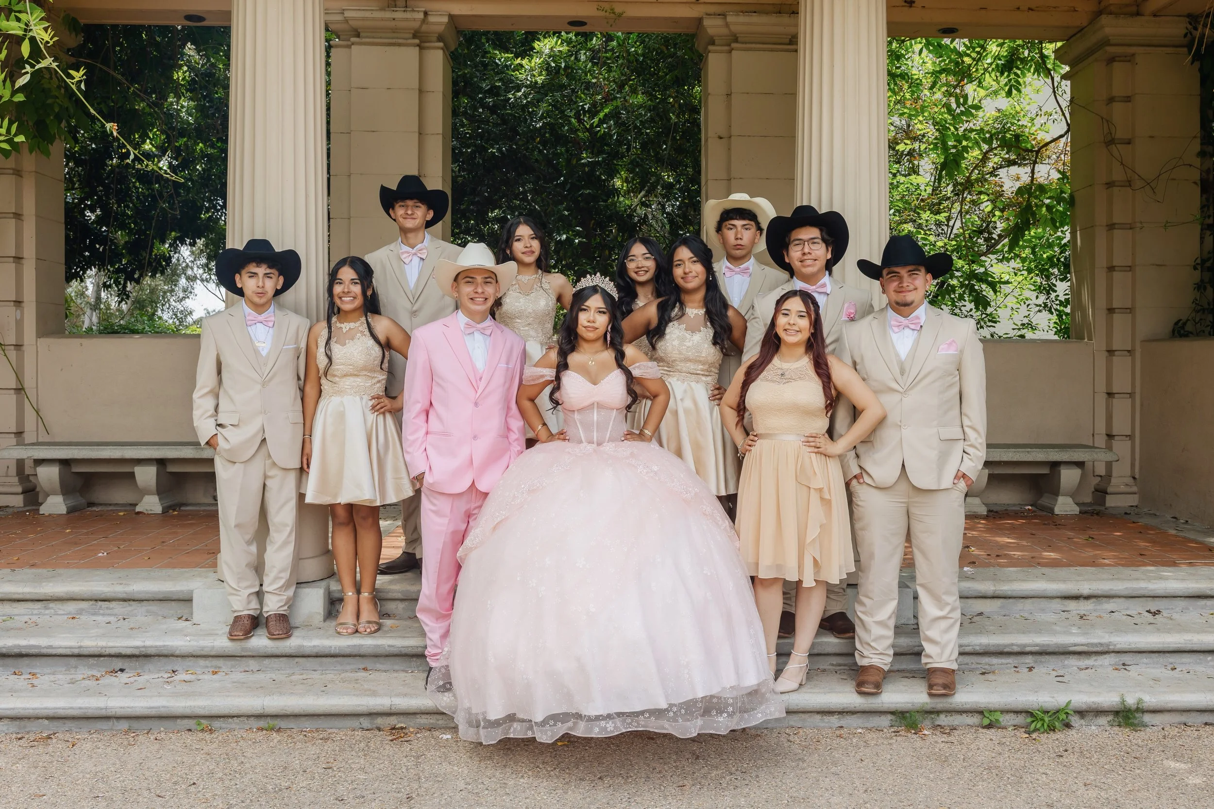 A group of young people dressed in formal attire, with some in beige suits and pink accessories, and a girl in a large pink ball gown, standing on steps in front of a classical pavilion with columns and greenery.