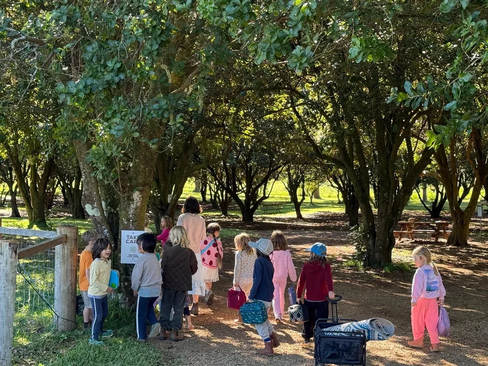 Children and adults standing in line under a large tree in a park, carrying suitcases and bags, with picnic tables and greenery in the background.