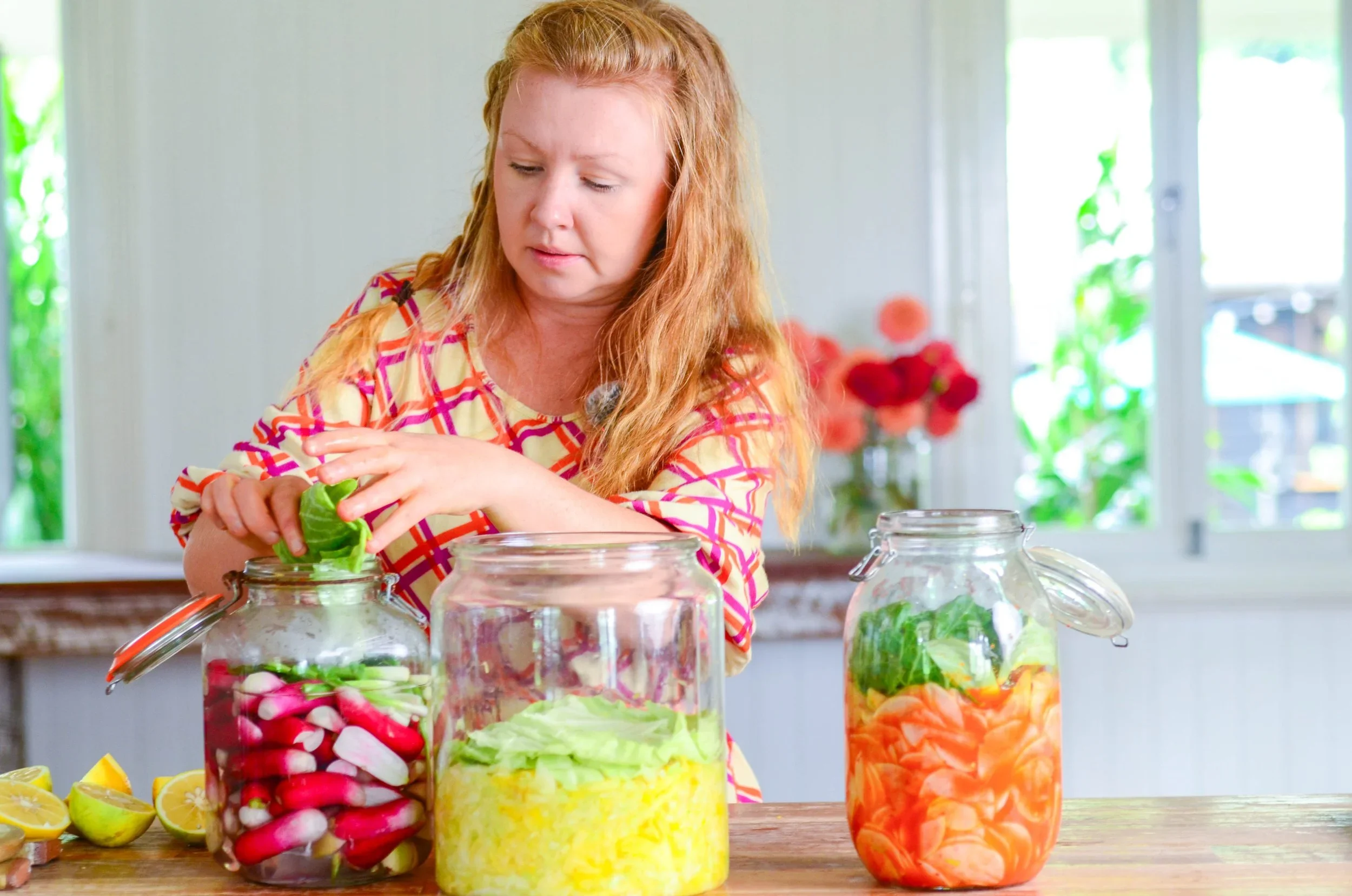 A woman with red hair in a colorful checkered shirt is preparing vegetables for pickling or preserving, surrounded by jars filled with sliced radishes, carrots, and leafy greens on a wooden table in a bright, sunny kitchen.