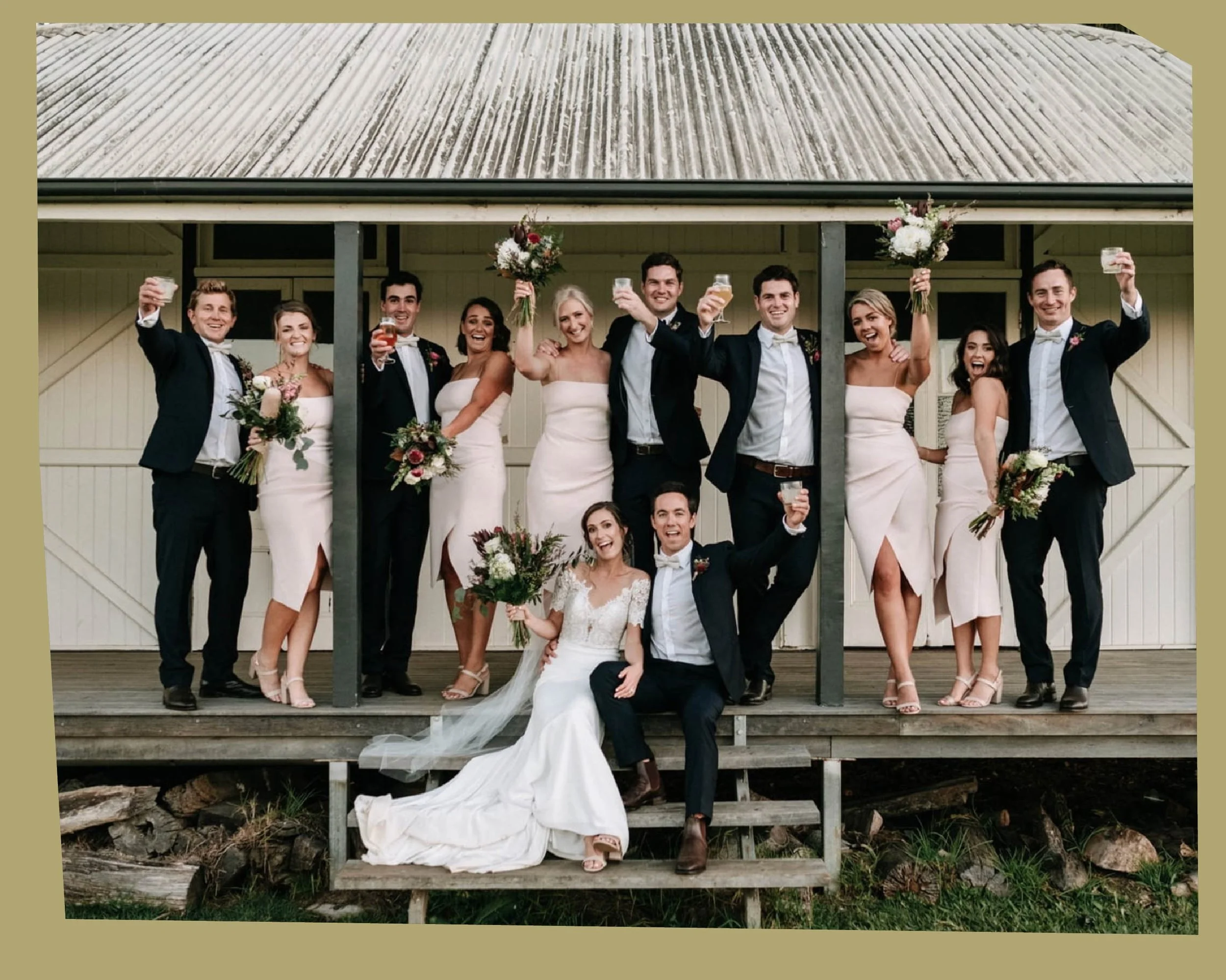 A group of wedding guests in formal attire celebrating on the porch of a house, raising glasses and holding bouquets, with a bride and groom seated at the front.