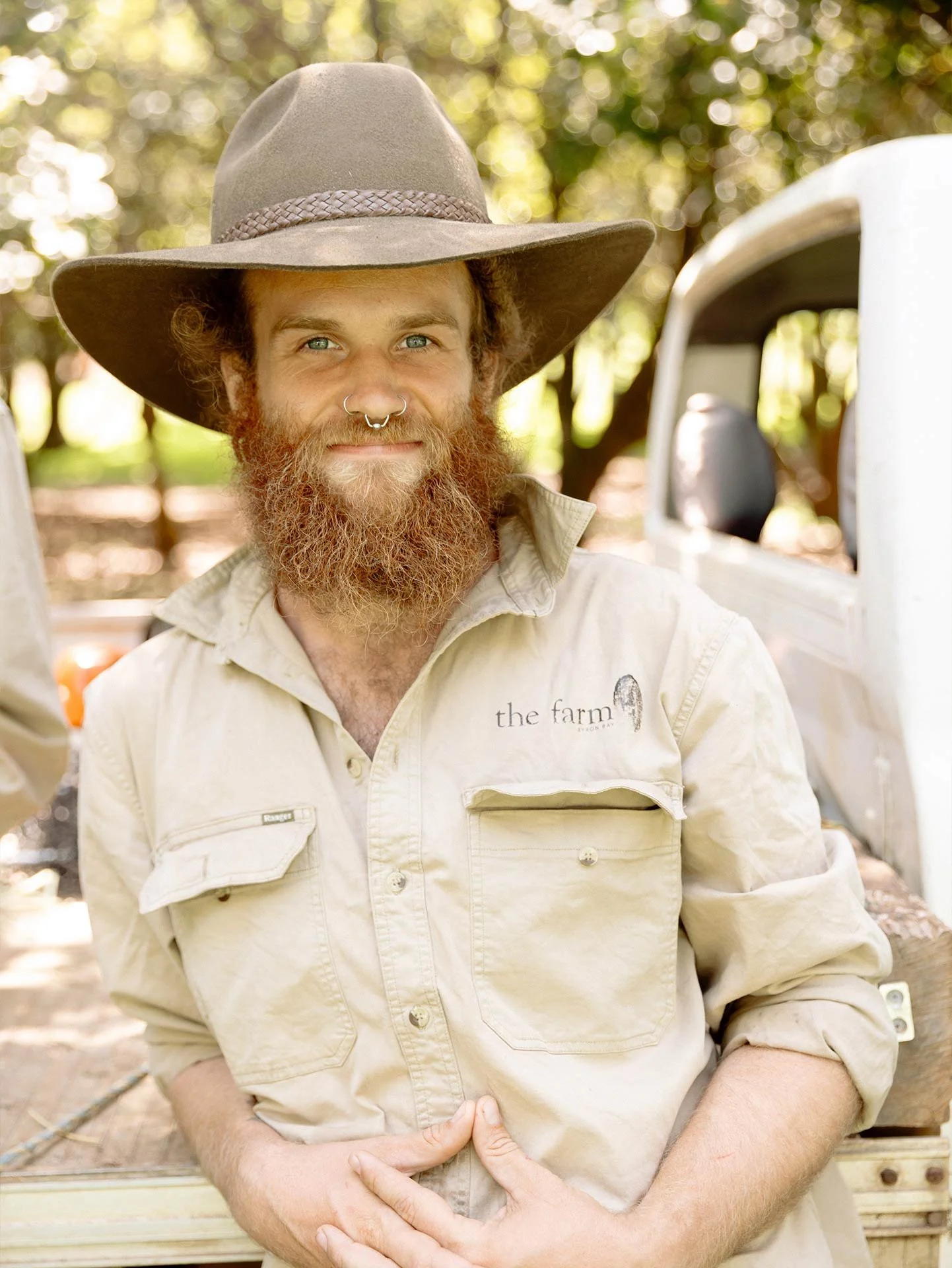 A man with a red beard and long hair, wearing a wide-brimmed hat and a beige shirt with 'the farm' embroidered on it, is outdoors. He has piercings on his nose and smile on his face, and is standing in front of a truck with trees in the background.