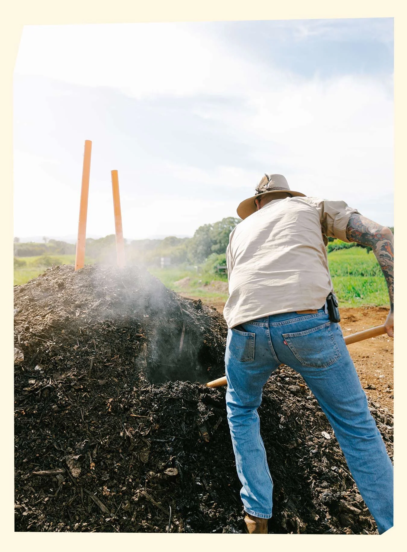 A person with tattoos wearing a hat and jeans is shoveling ash or charred wood from a large fire or compost pile outdoors under a partly cloudy sky.
