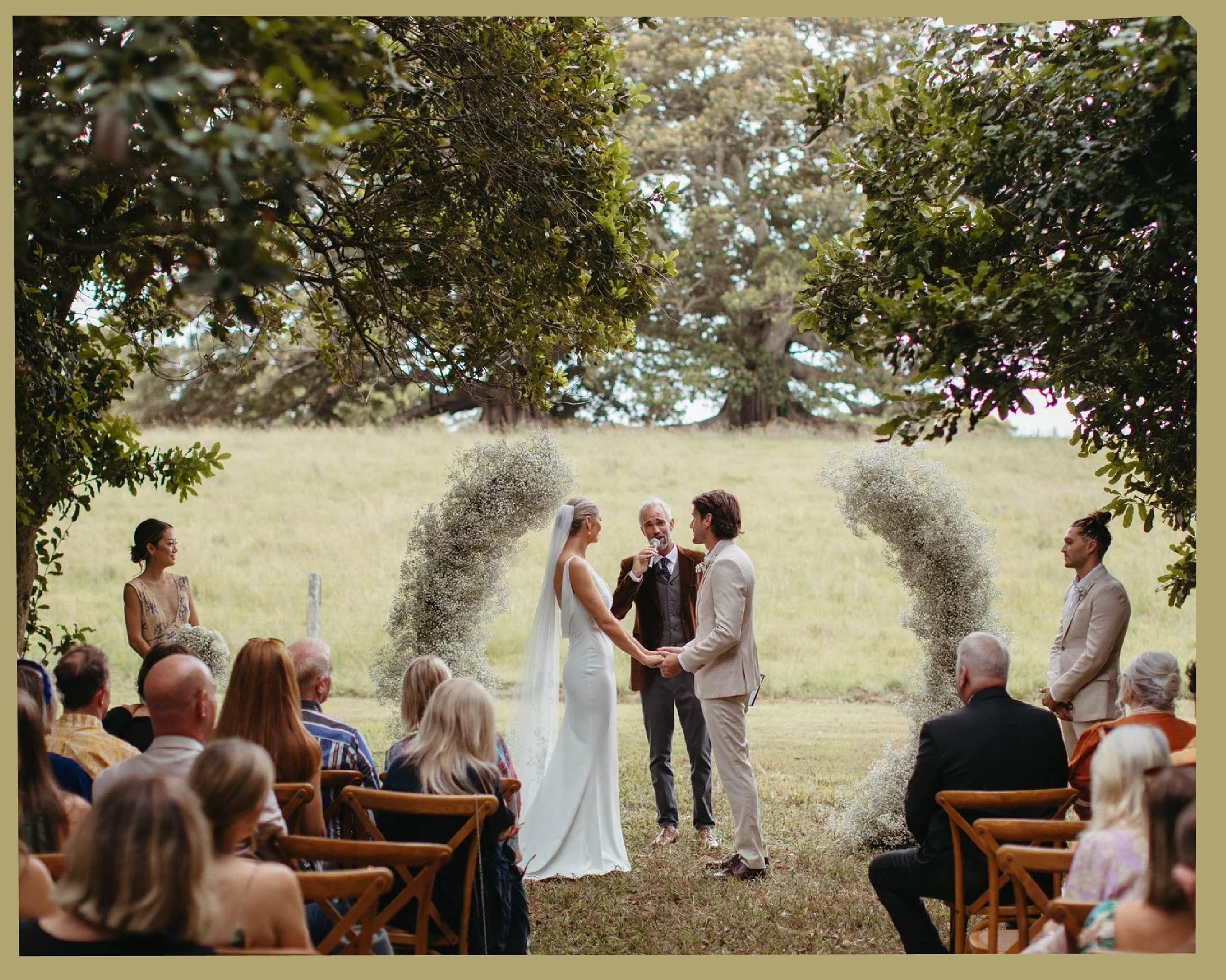 A wedding ceremony outdoors in a grassy field, with the bride and groom exchanging vows under a canopy of trees, surrounded by guests seated on chairs, and a officiant conducting the ceremony.