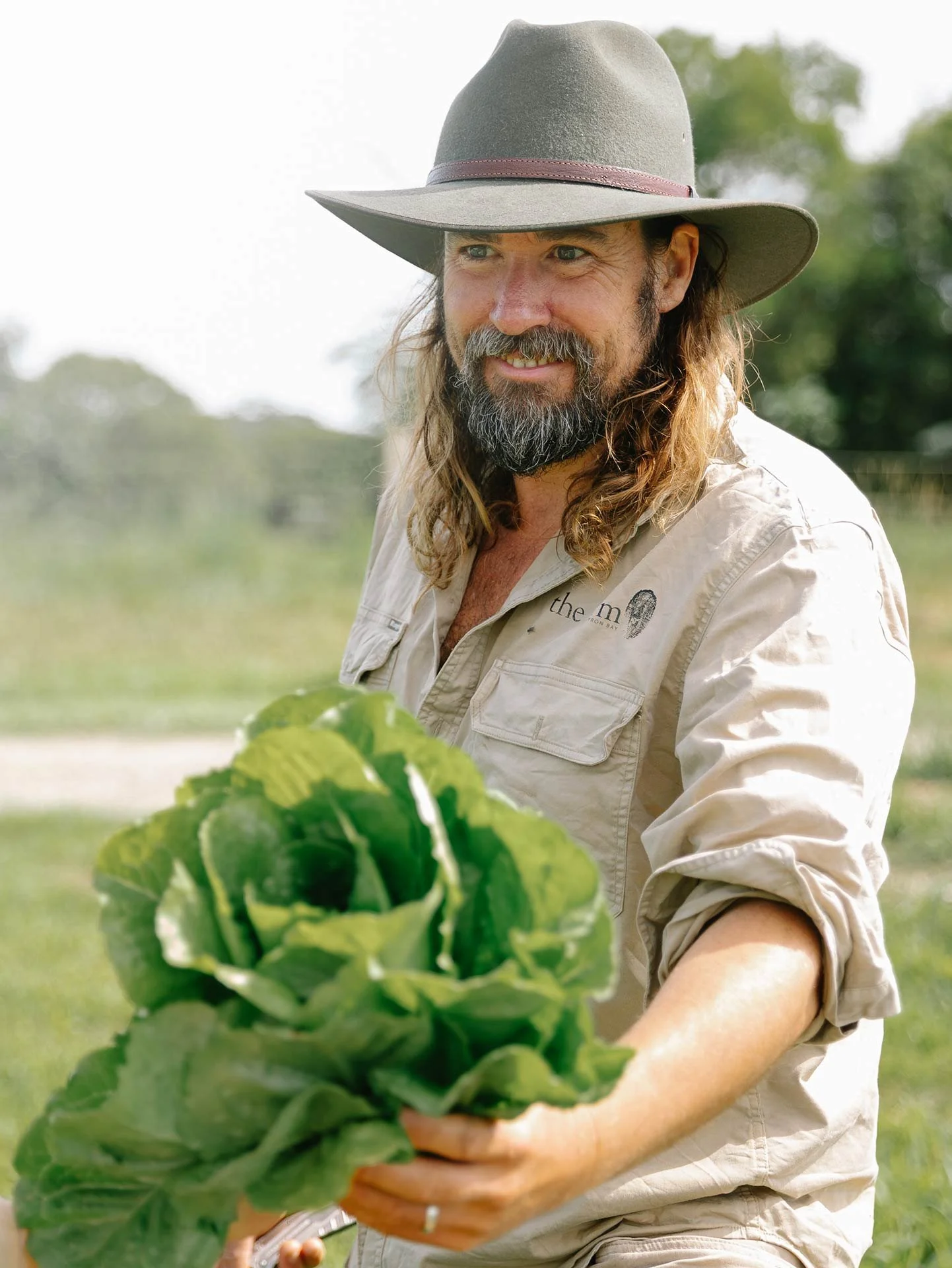 A man with long hair and a beard wearing a wide-brimmed hat and a beige shirt holding fresh green lettuce in an outdoor setting with trees and grass.