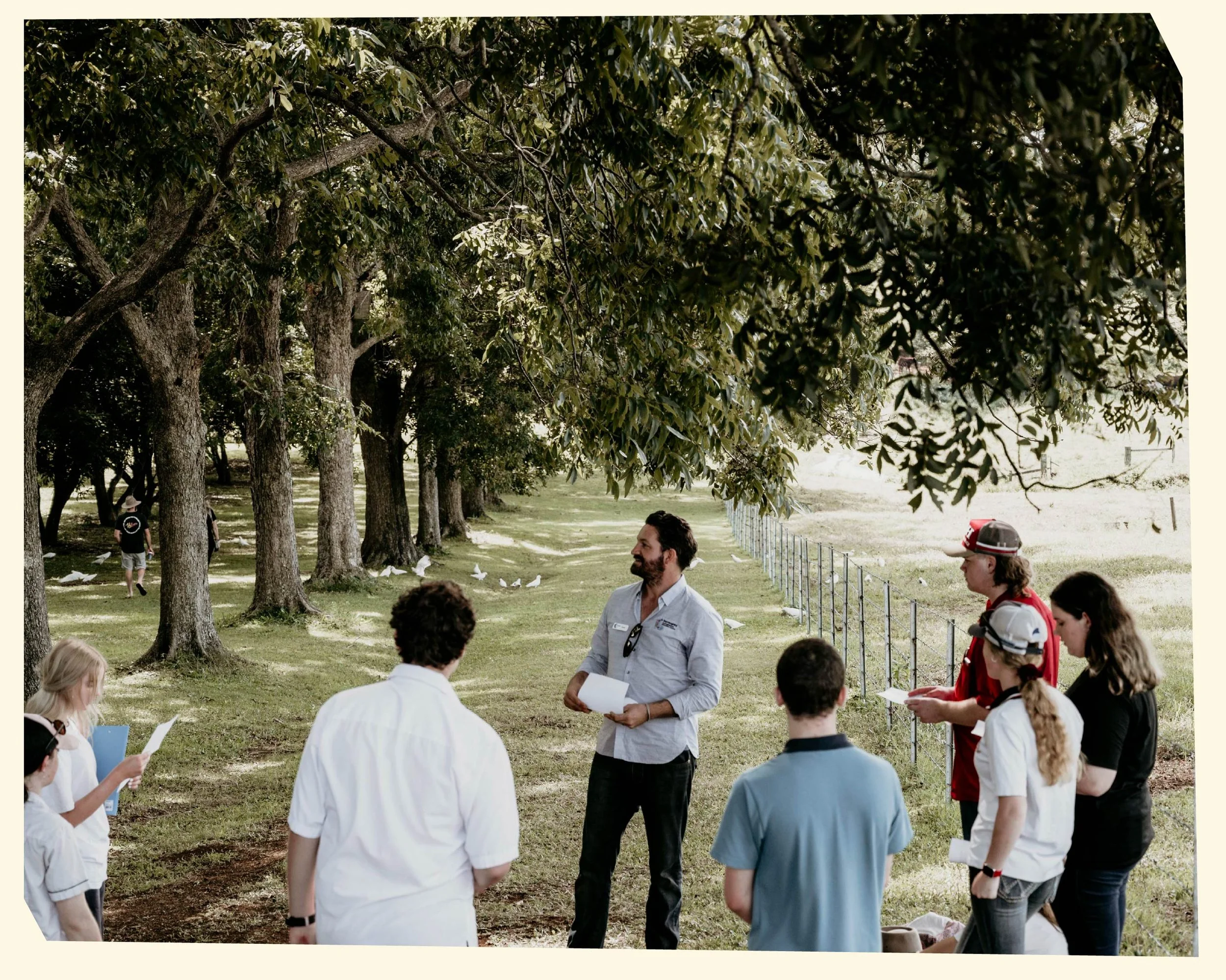 People are gathered outdoors in a park under trees, participating in a group activity or discussion. Several individuals are holding papers, and a man appears to be leading or speaking to the group.