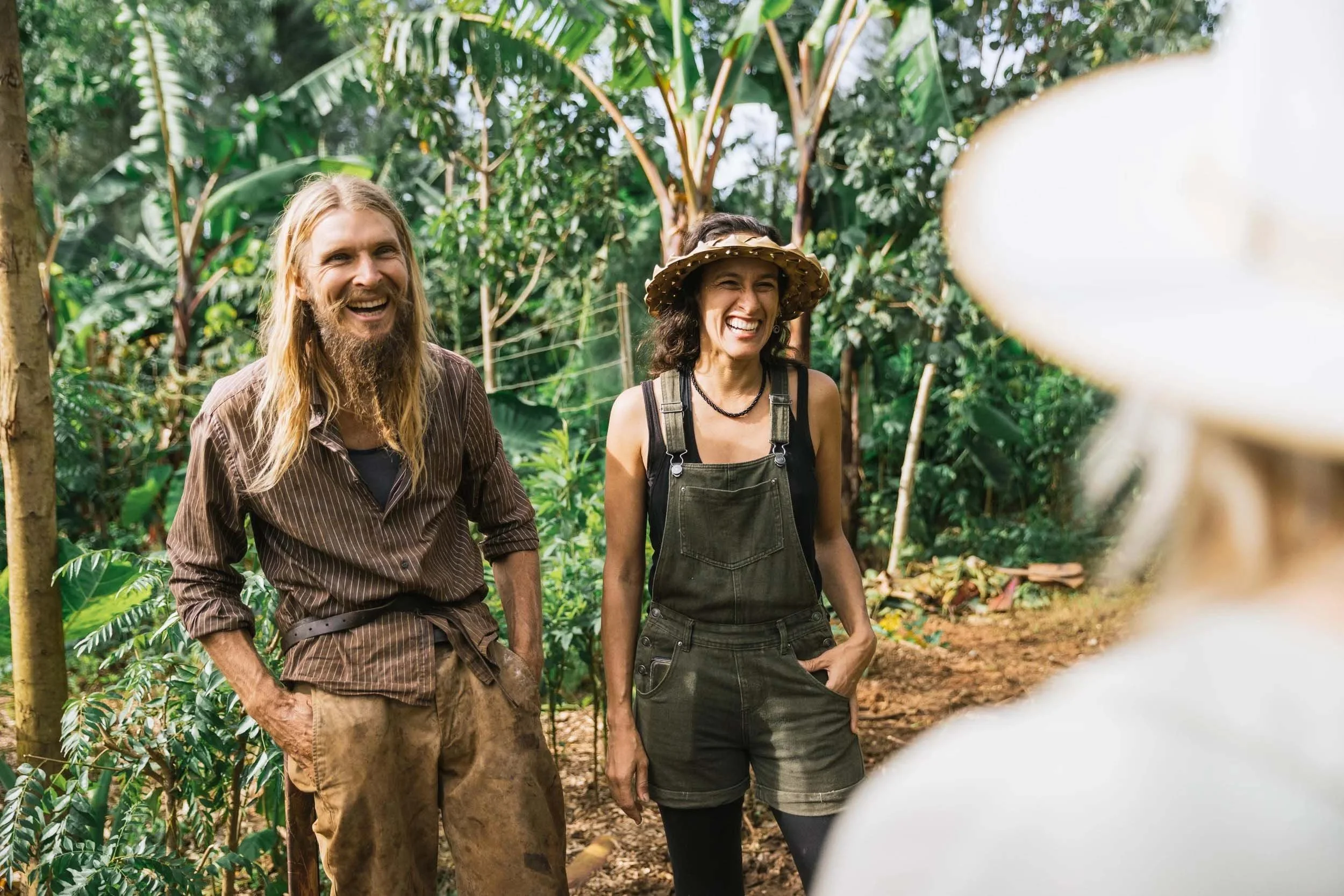 Two people smiling and walking through a lush, green jungle or forest, with the photo taken from a blurred foreground object.