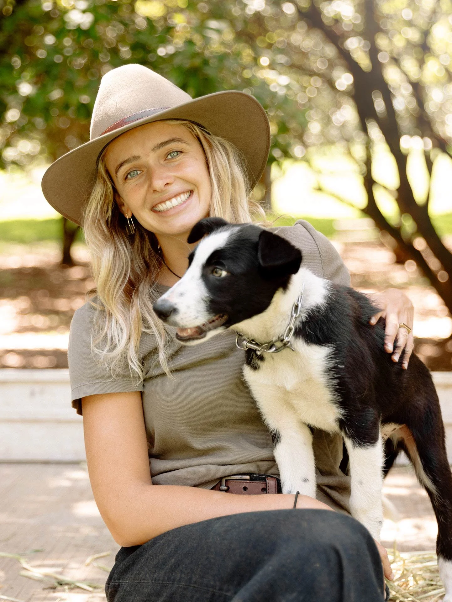 A young woman with blonde hair, wearing a beige wide-brimmed hat and a gray t-shirt, smiling while holding a black and white Border Collie puppy outdoors with a blurred background of trees and sunlight.